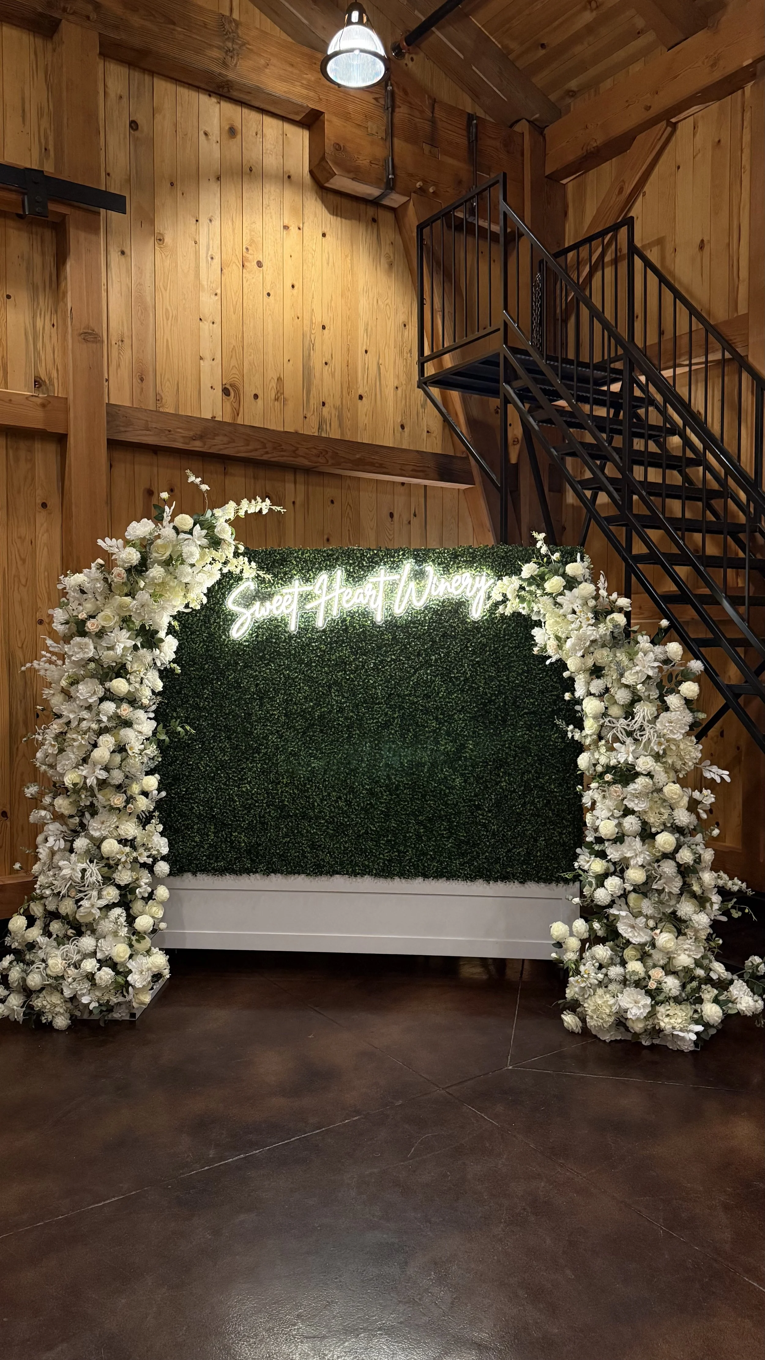 Flower wall with white flowers and greenery, and a neon sign that reads "Sweet Heart Winery" in cursive writing.