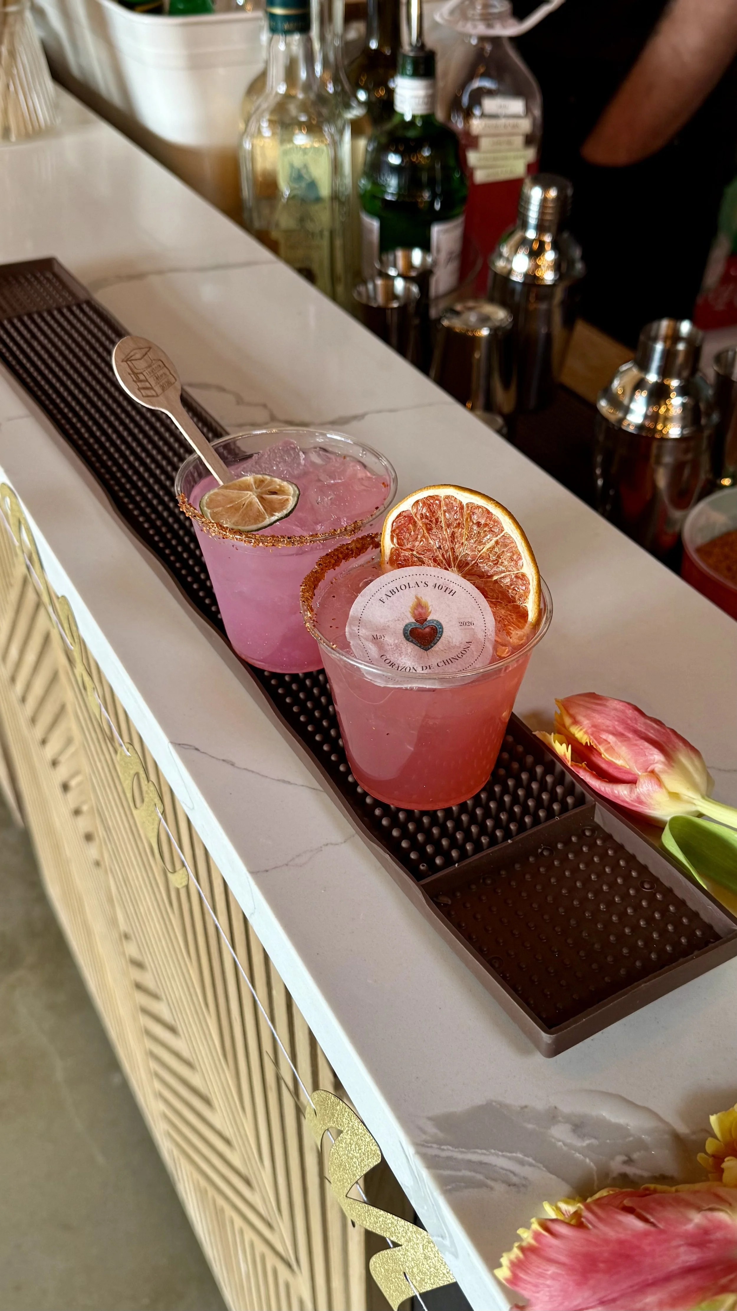 Two pink cocktails garnished with dried citrus slices, one with a small lemon slice and the other with a circular label, on a bar counter with liquor bottles and festive decorations in the background.