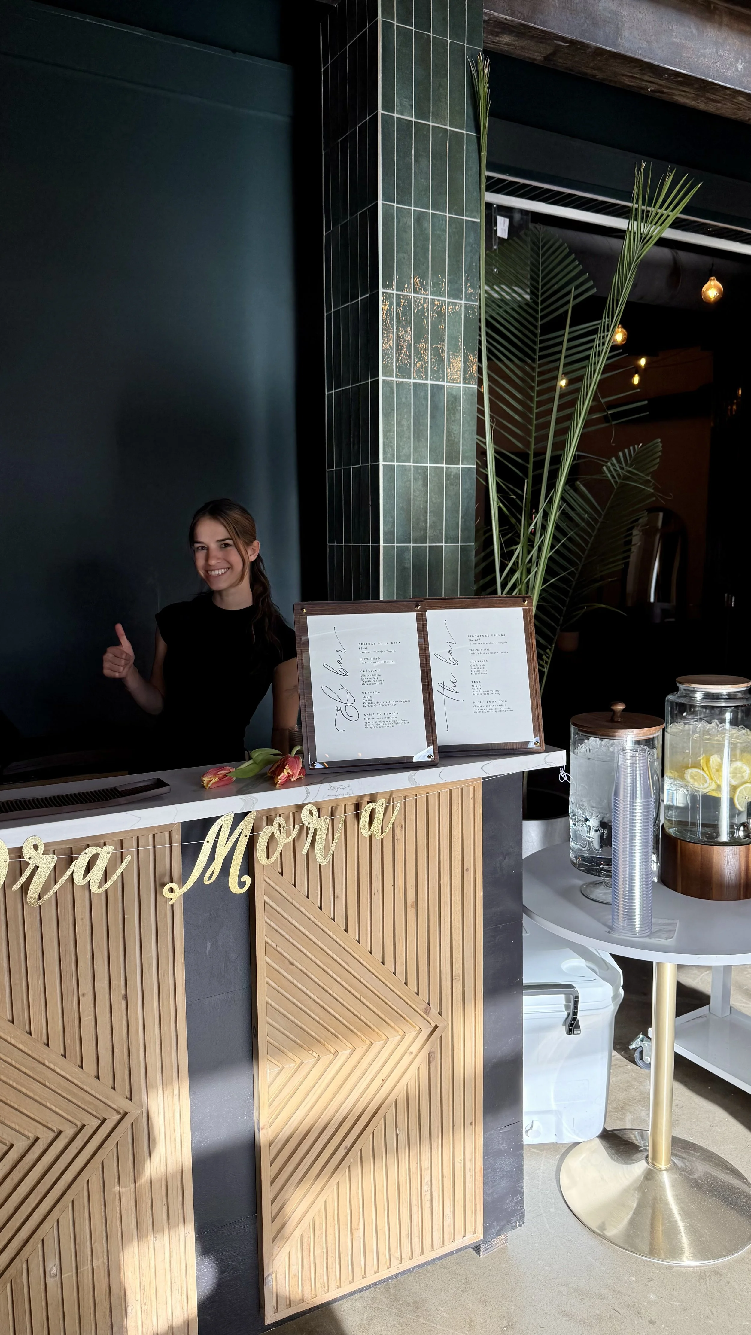 A smiling woman giving a thumbs up behind a wooden counter with a black menu board and a large green plant, in a modern, stylish indoor setting with dark walls and warm lighting.