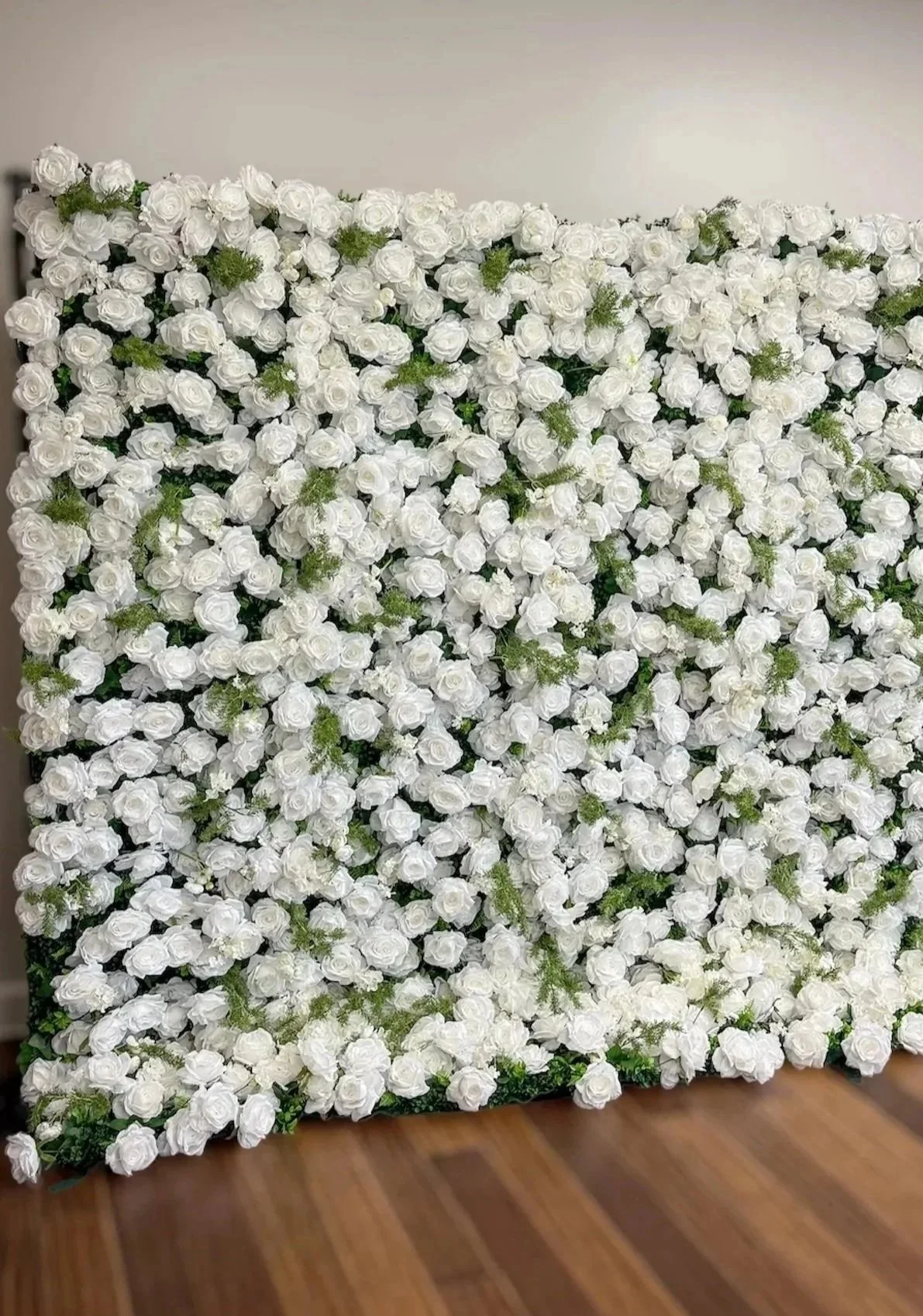 A large rectangular arrangement of white roses with some green foliage, set against a plain light-colored wall and resting on a wooden floor.