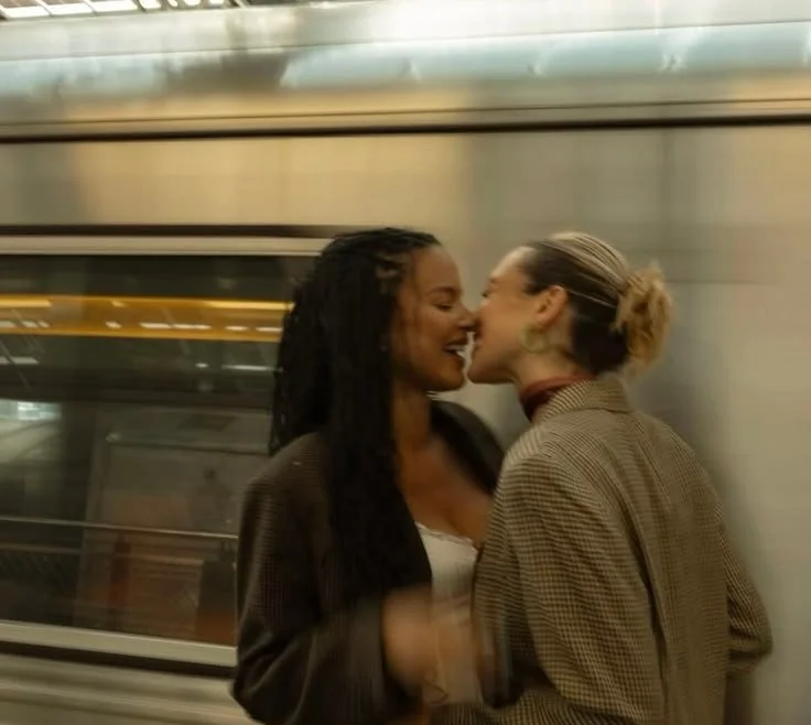 Two women sharing an intimate moment on a subway train, close to each other with noses touching, smiling, and dressed in casual clothing.