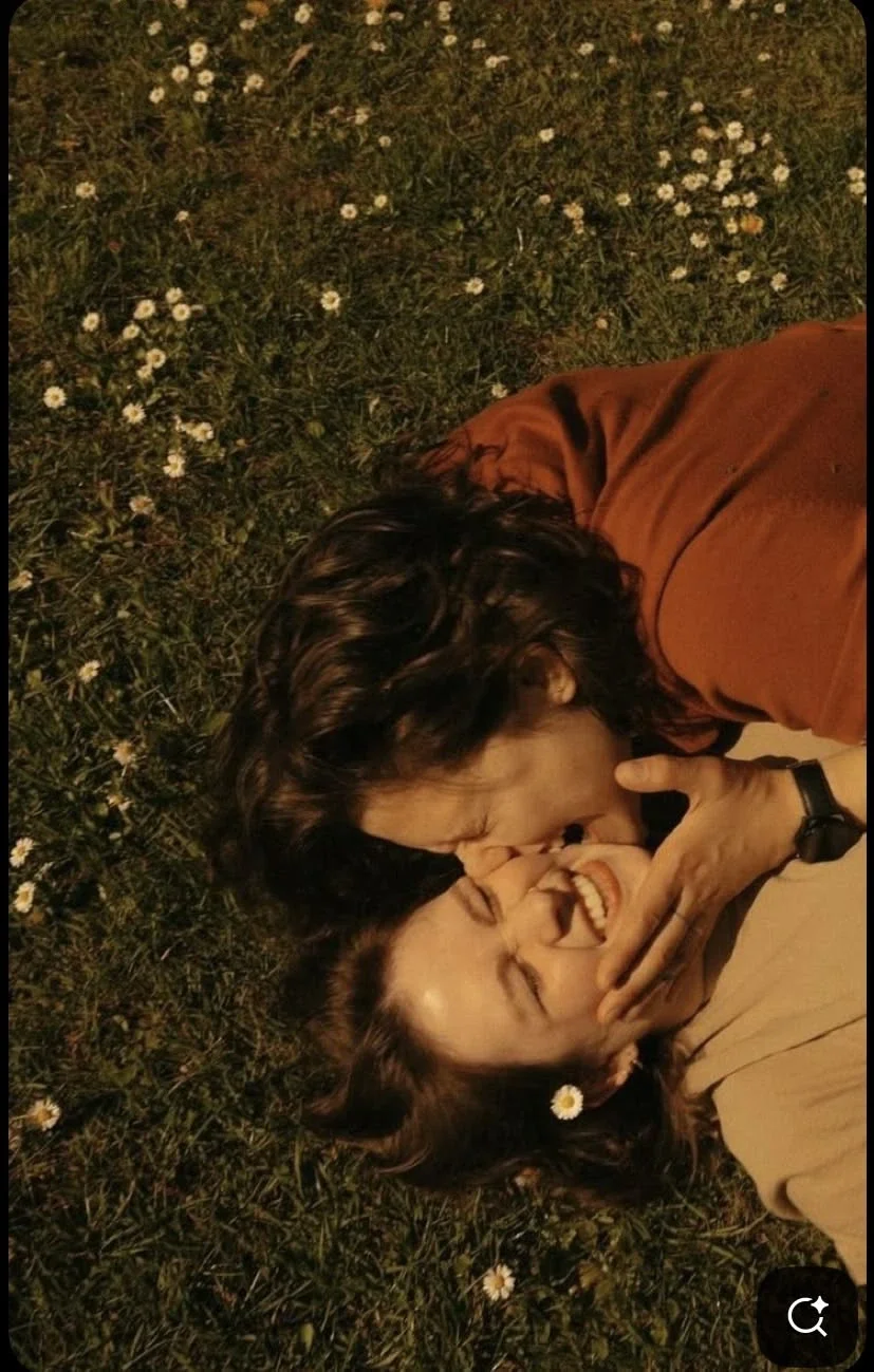 Two young women lying on grass surrounded by small white daisies, playfully covering each other's faces and smiling.