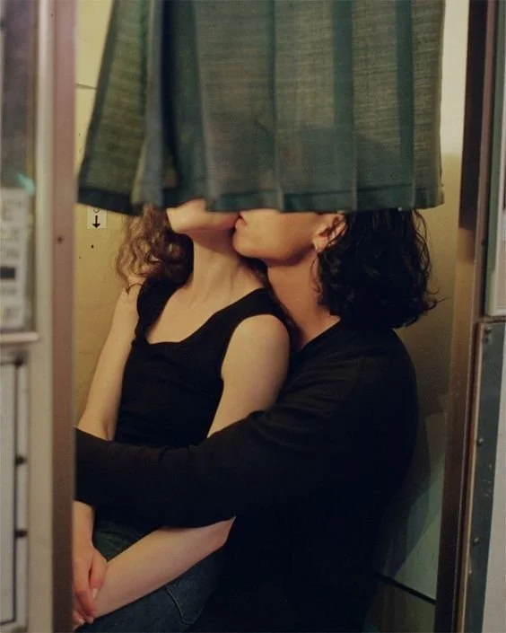 Two people are sitting close together with their faces hidden behind a hanging curtain, creating a mirror-like reflection.