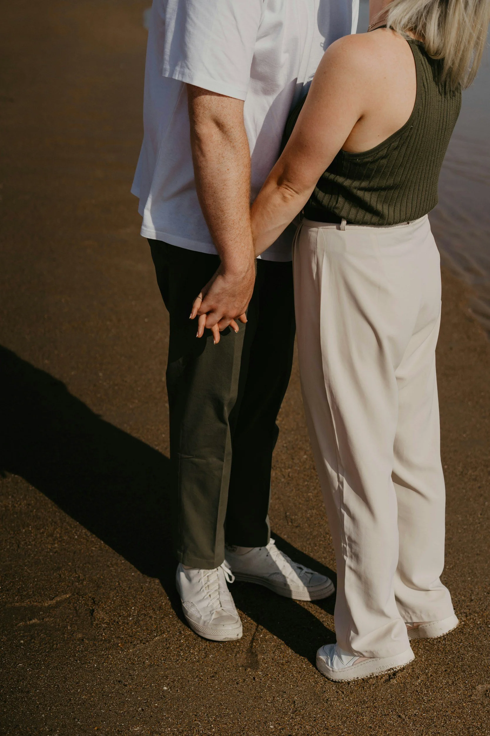A couple holding hands on a sandy beach, facing each other. The woman is wearing a green sleeveless top and white pants, while the man is in a white shirt, dark pants, and white sneakers.