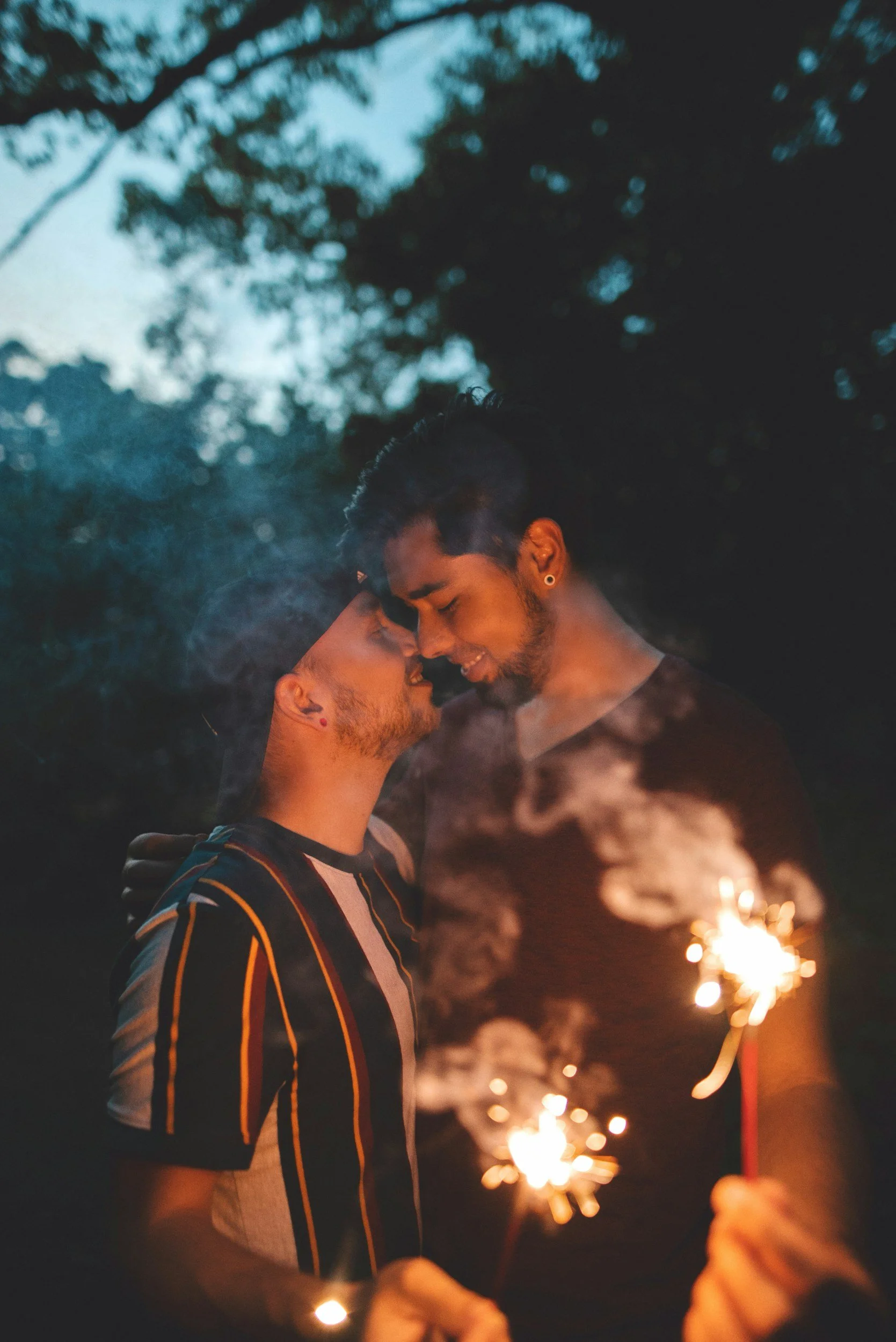 Two men embracing each other at dusk, holding sparklers, with trees in the background.