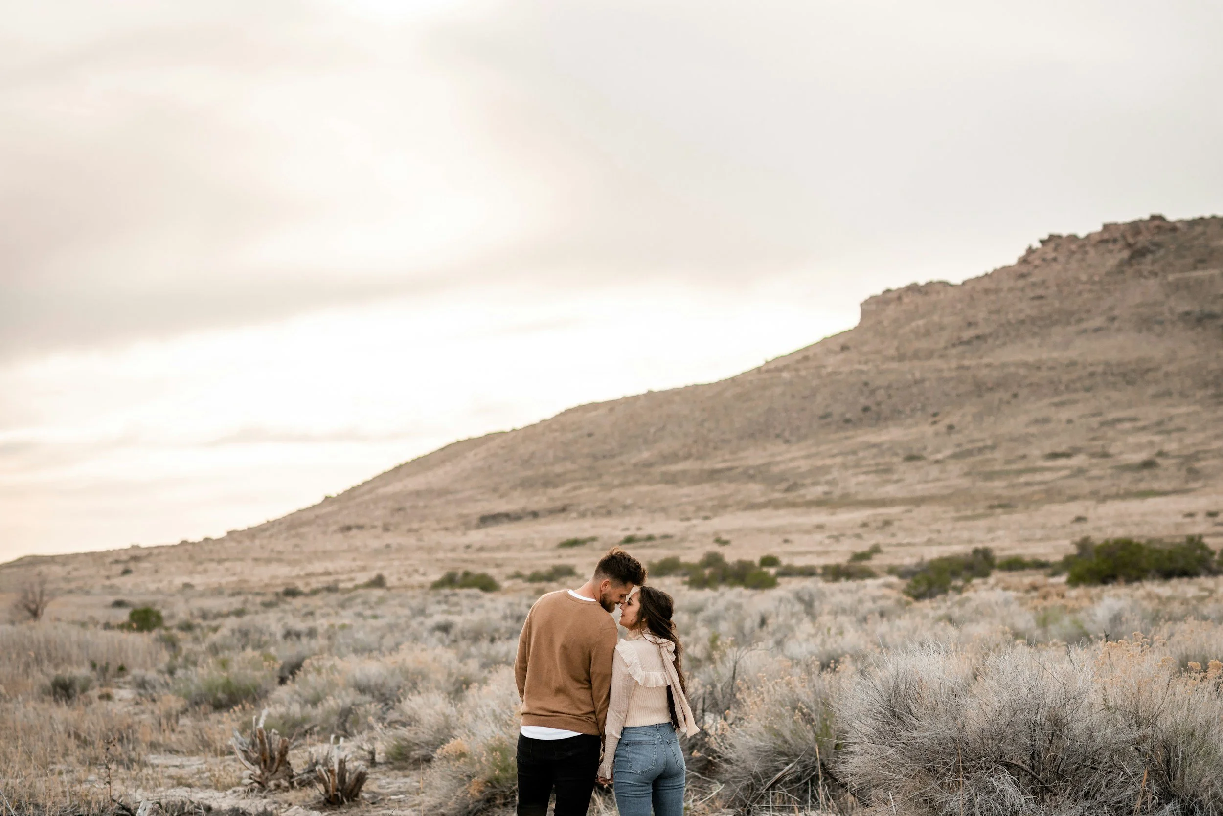 A couple standing close together in a desert landscape with sparse bushes and a mountain in the background.