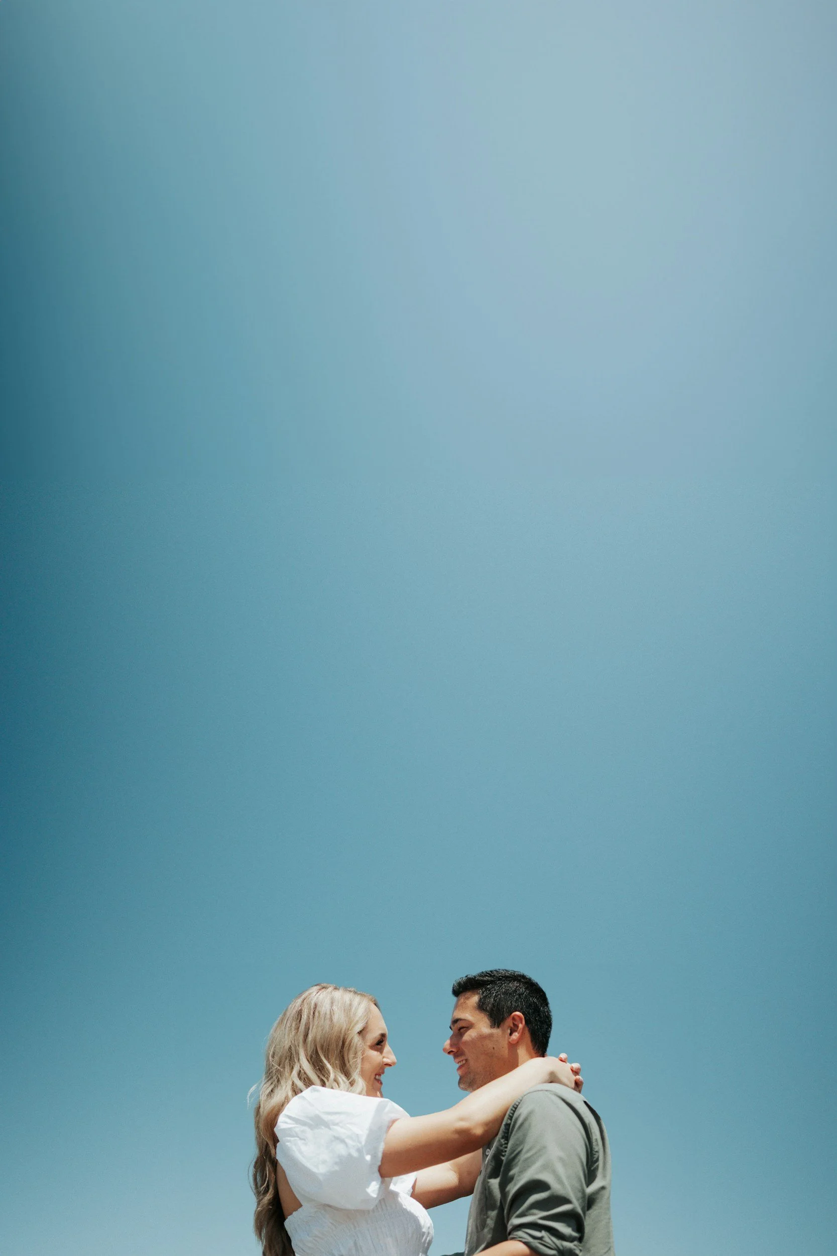 A couple smiling and embracing outdoors under a bright blue sky.