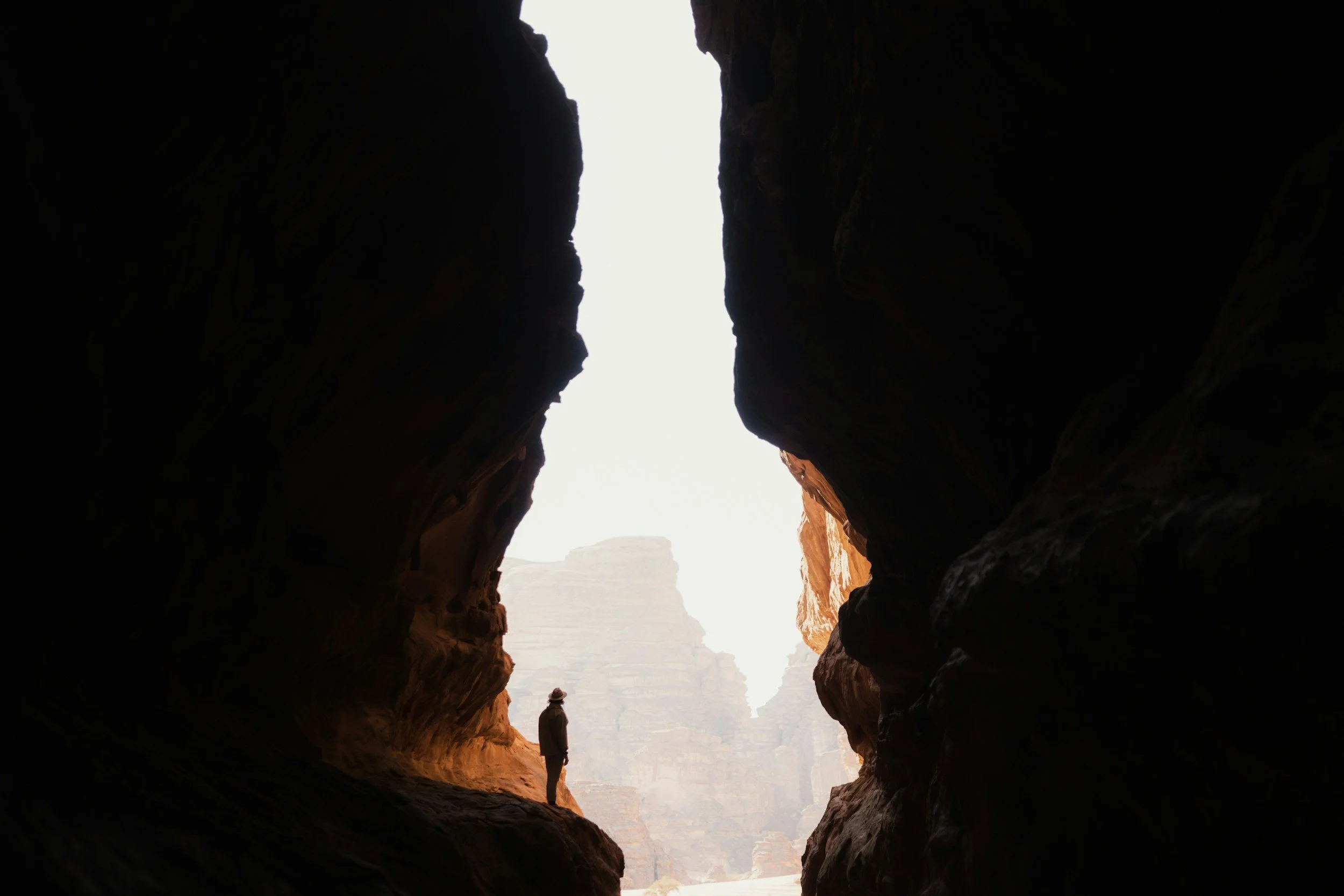 A person wearing a hat standing inside a dark canyon, looking out at a bright sky and distant rock formations.