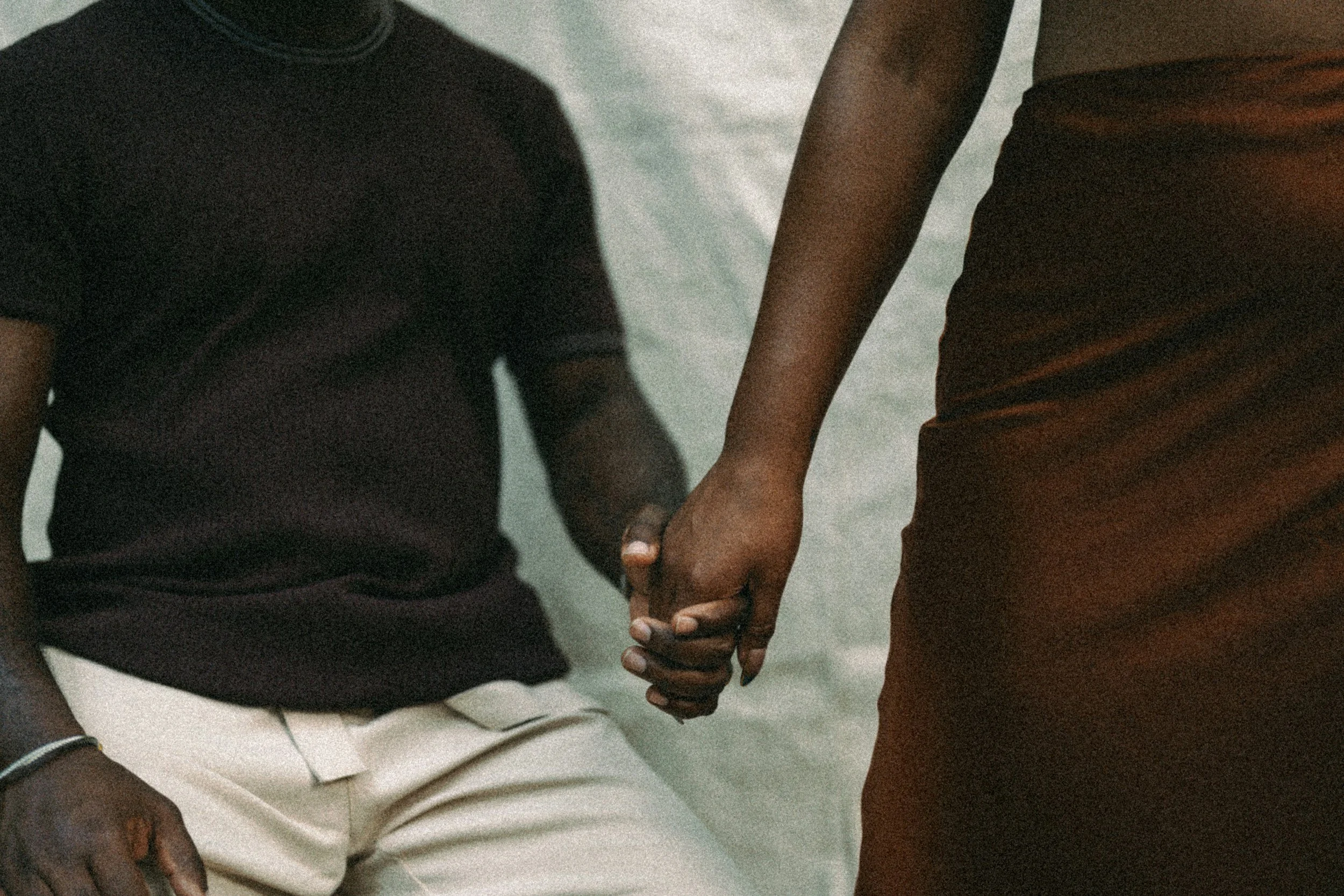 Close-up of two people holding hands, one wearing a black t-shirt and beige pants, and the other wearing an orange-brown shirt and matching pants.