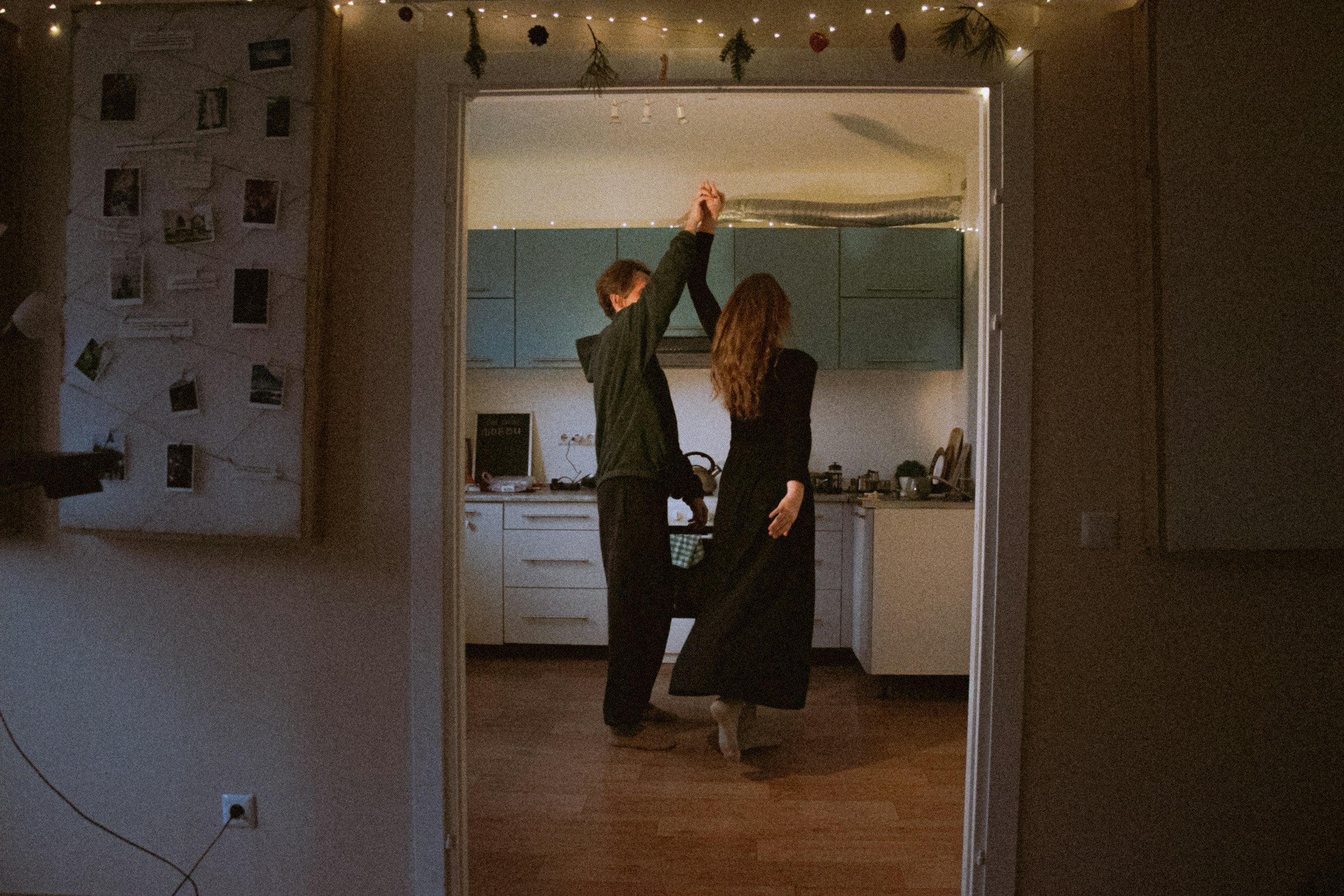 A couple dancing in a kitchen with string lights hanging from the ceiling, viewed through a doorway.