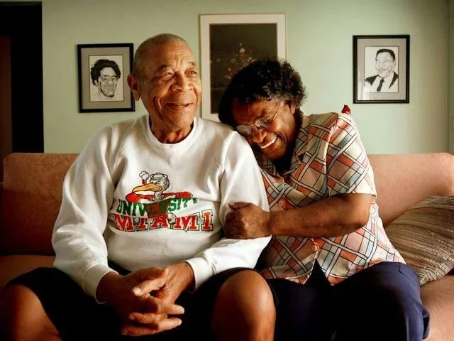 Two people sitting on a sofa, smiling and laughing together. One is an older man in a white sweatshirt, and the other is a woman in a plaid shirt. There are black-and-white portraits hanging on the wall behind them.