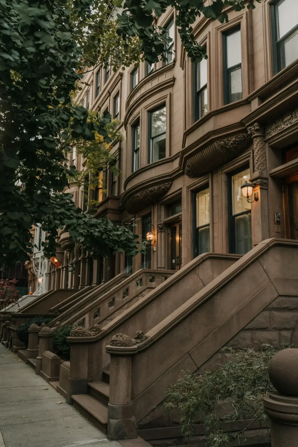 Brownstone row house with elaborate stonework, staircases, and ornate lampposts on the front steps. Green trees partially cover the building, creating a classic city street scene.