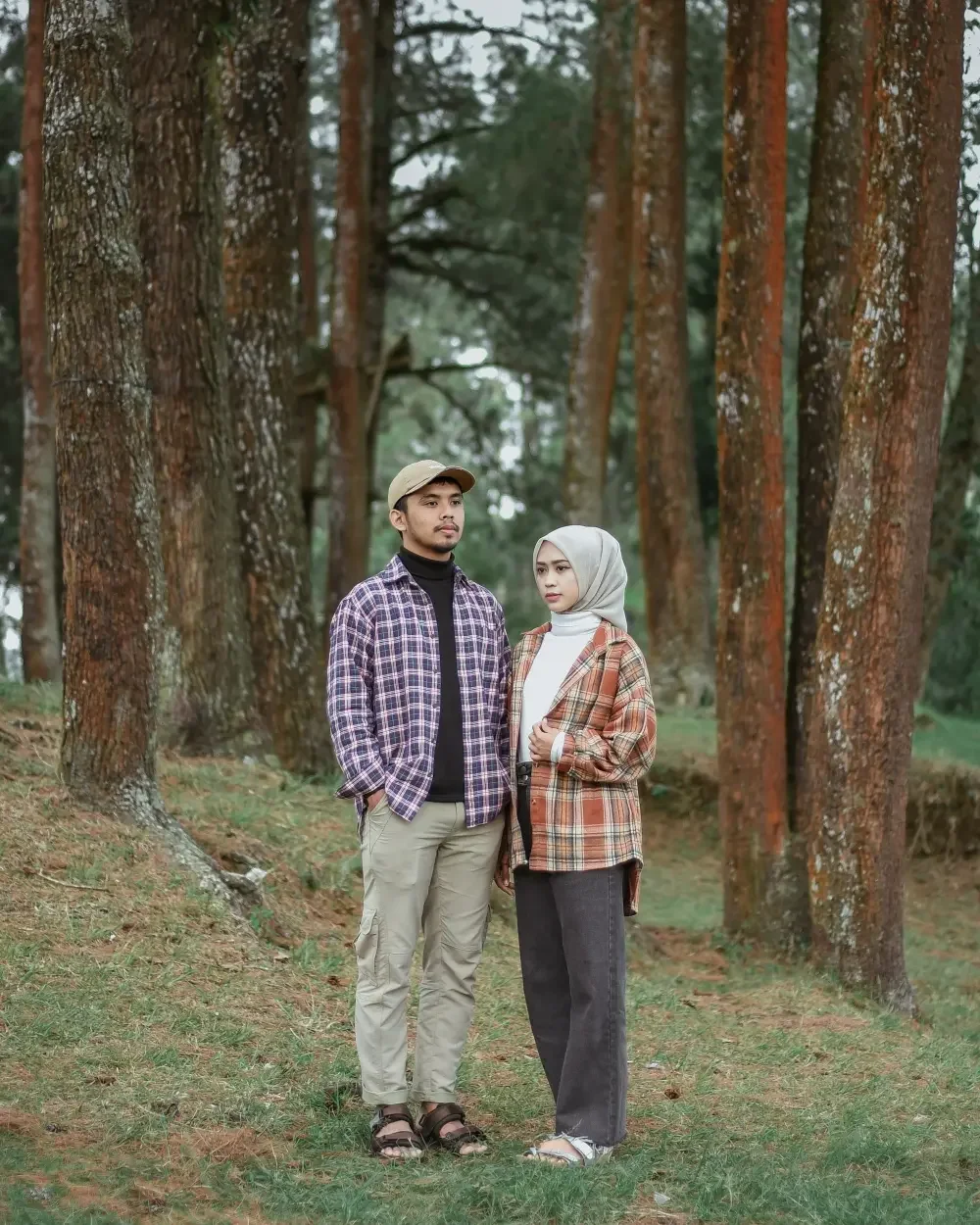 A man and woman standing together in a forest with tall trees in the background. The man is wearing a beige cap, checkered shirt, black turtleneck, and beige cargo pants. The woman is wearing a gray hijab, checkered jacket, white top, and dark jeans. They appear to be posing for a photo outdoors.