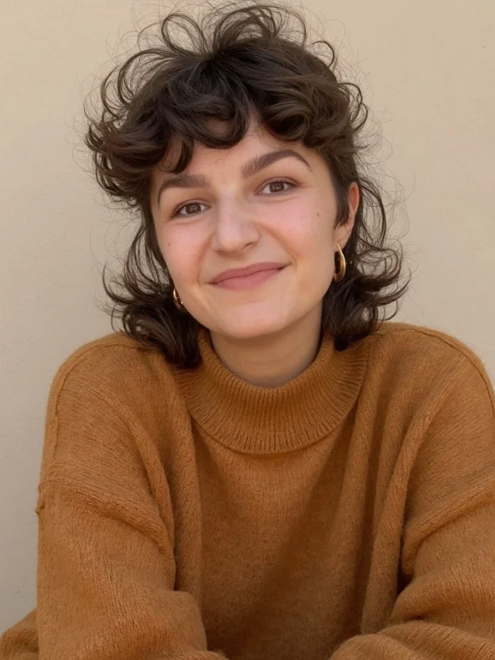 A young woman with curly brown hair wearing a brown turtleneck sweater and gold hoop earrings, smiling at the camera with a plain light-colored background.