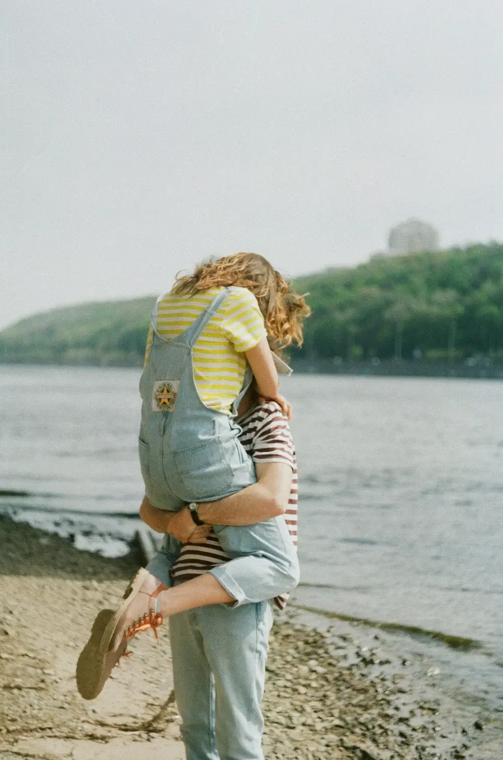 A man lifting a girl in front of a body of water with a green hill in the background.