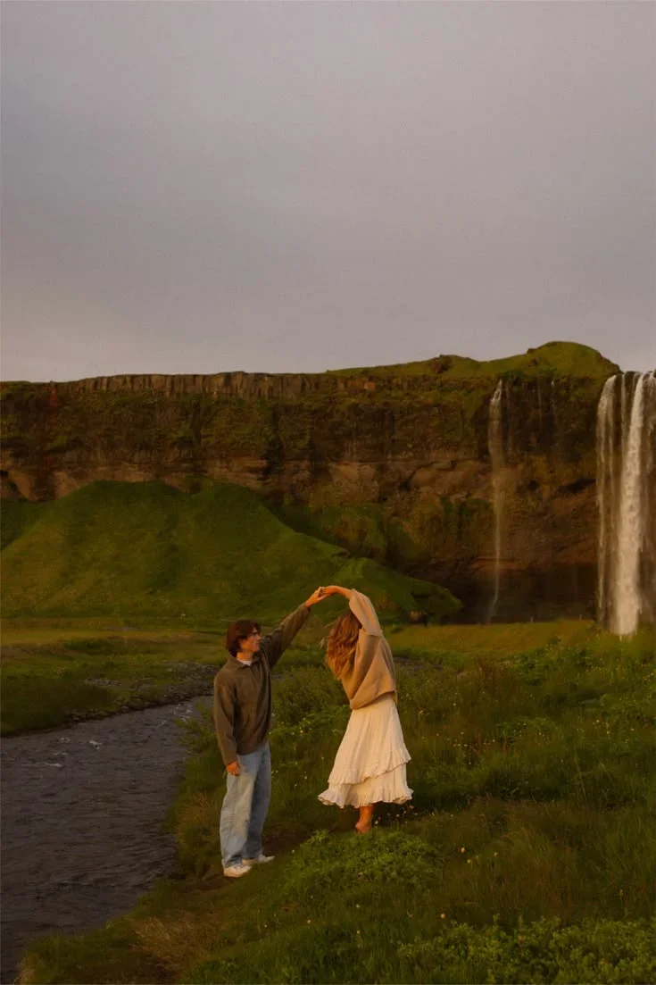 A couple dancing on a grassy area near a river with a waterfall and green cliffs in the background during sunset.