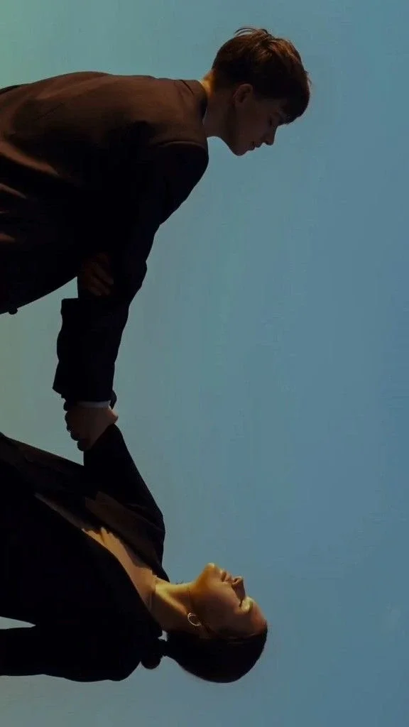A man leaning over and looking down at a woman who is lying on the ground, both wearing black clothing, against a plain light blue background.