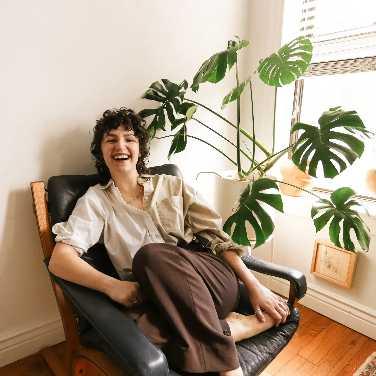 A woman with curly dark hair, wearing a beige shirt and brown pants, sitting on a black leather chair, smiling. Behind her is a large green monstera plant near a window with white blinds, and framed artwork on the floor.