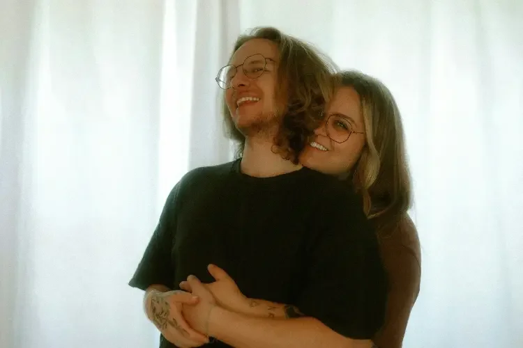 Two people, a man with glasses and wavy hair, and a woman with glasses and straight hair, smiling and hugging in front of a white curtain.