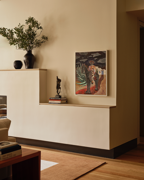 Interior of a living room with a curved beige wall, a built-in white shelf holding books and decorative items, a large potted plant in a black vase, and a colorful painting on the wall. A wooden coffee table with books and a rug are partially visible