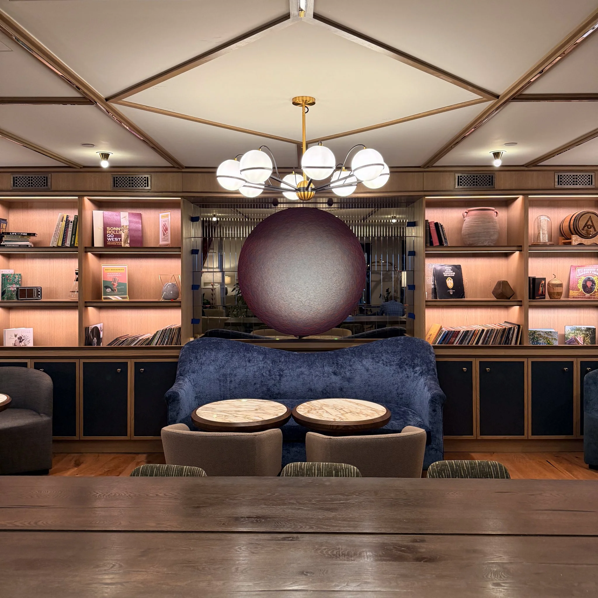 Interior of a cozy lounge or library with built-in wooden shelves filled with books, decorative items, and vinyl records. In front, there is a dark blue velvet sofa with two small round marble-top coffee tables, and a large spherical hanging art piec