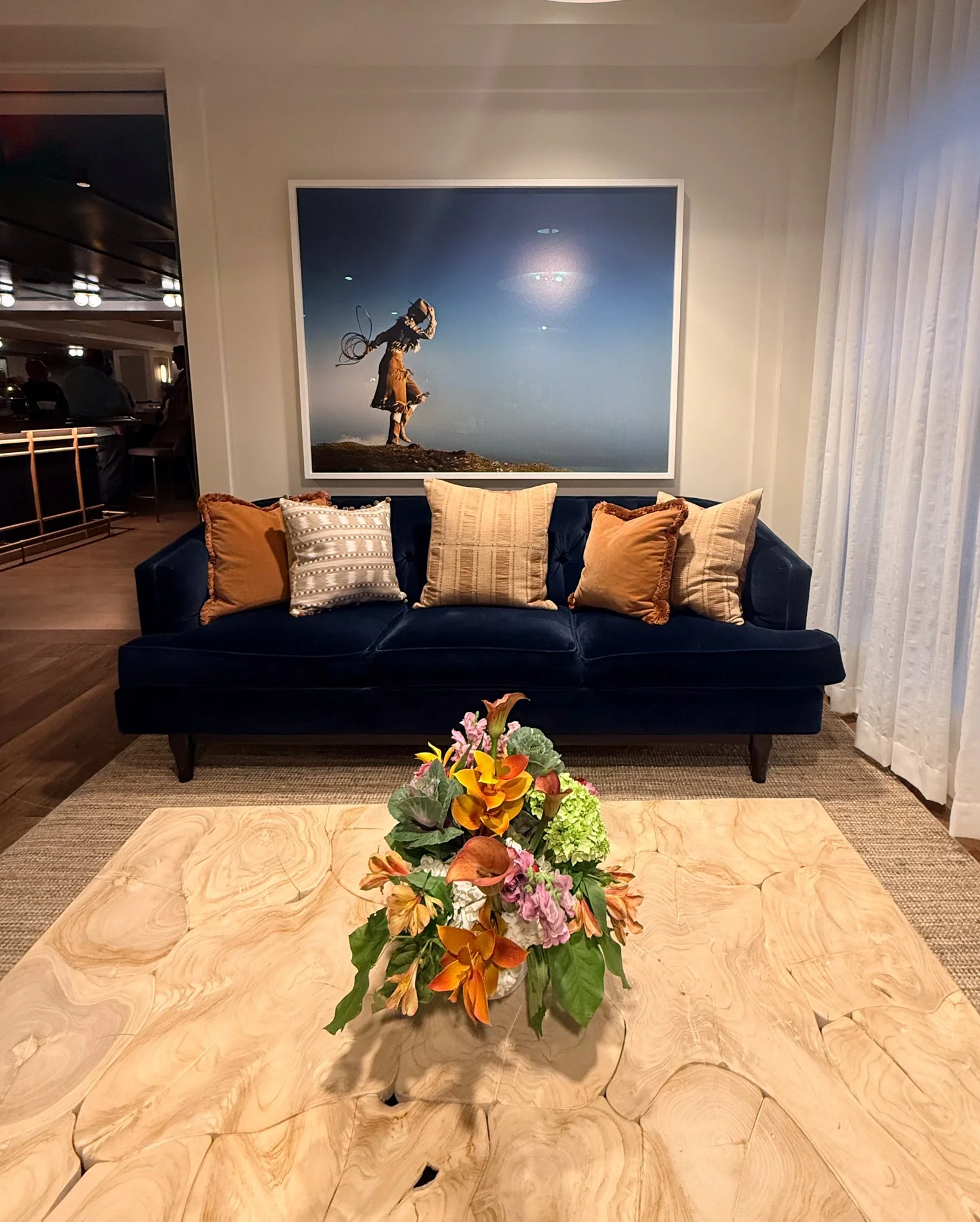 A hotel lobby or living area with a navy blue sofa with five pillows in shades of gold, beige, and silver, a wooden coffee table with a floral arrangement, large framed artwork of a child with a kite on the wall behind, and sheer white curtains on th