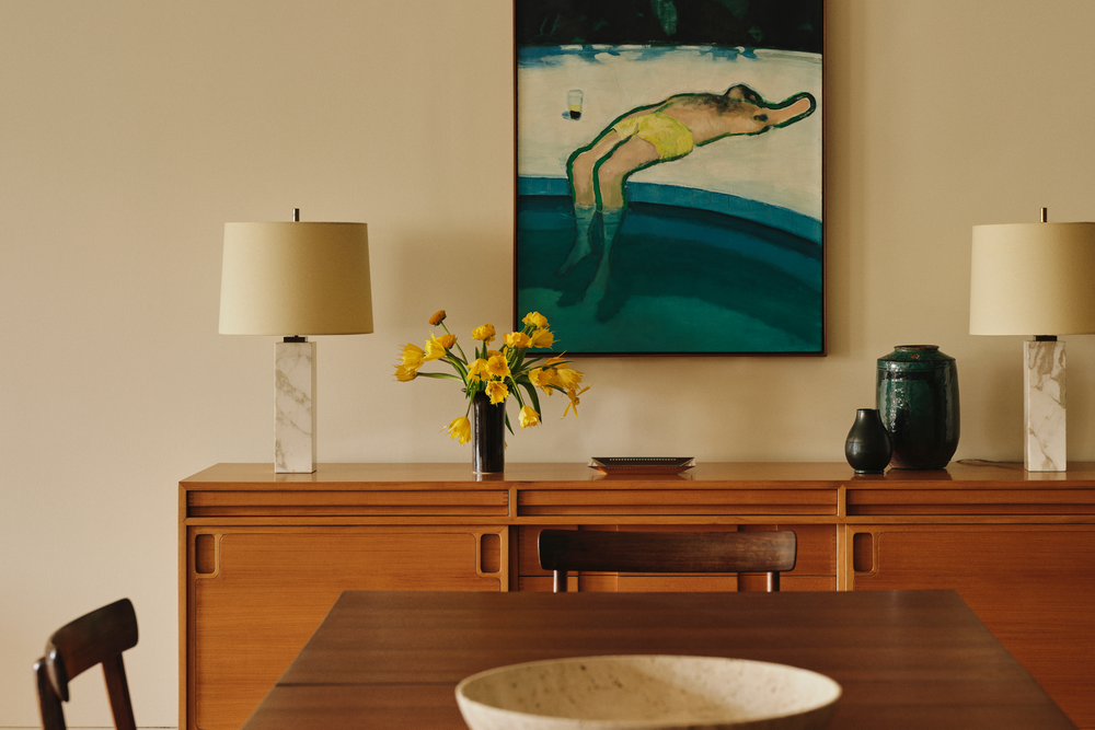 Interior of a dining room with a wooden sideboard, two lamps, a vase with yellow flowers, and abstract painting of a reclining person in yellow shorts on the wall.