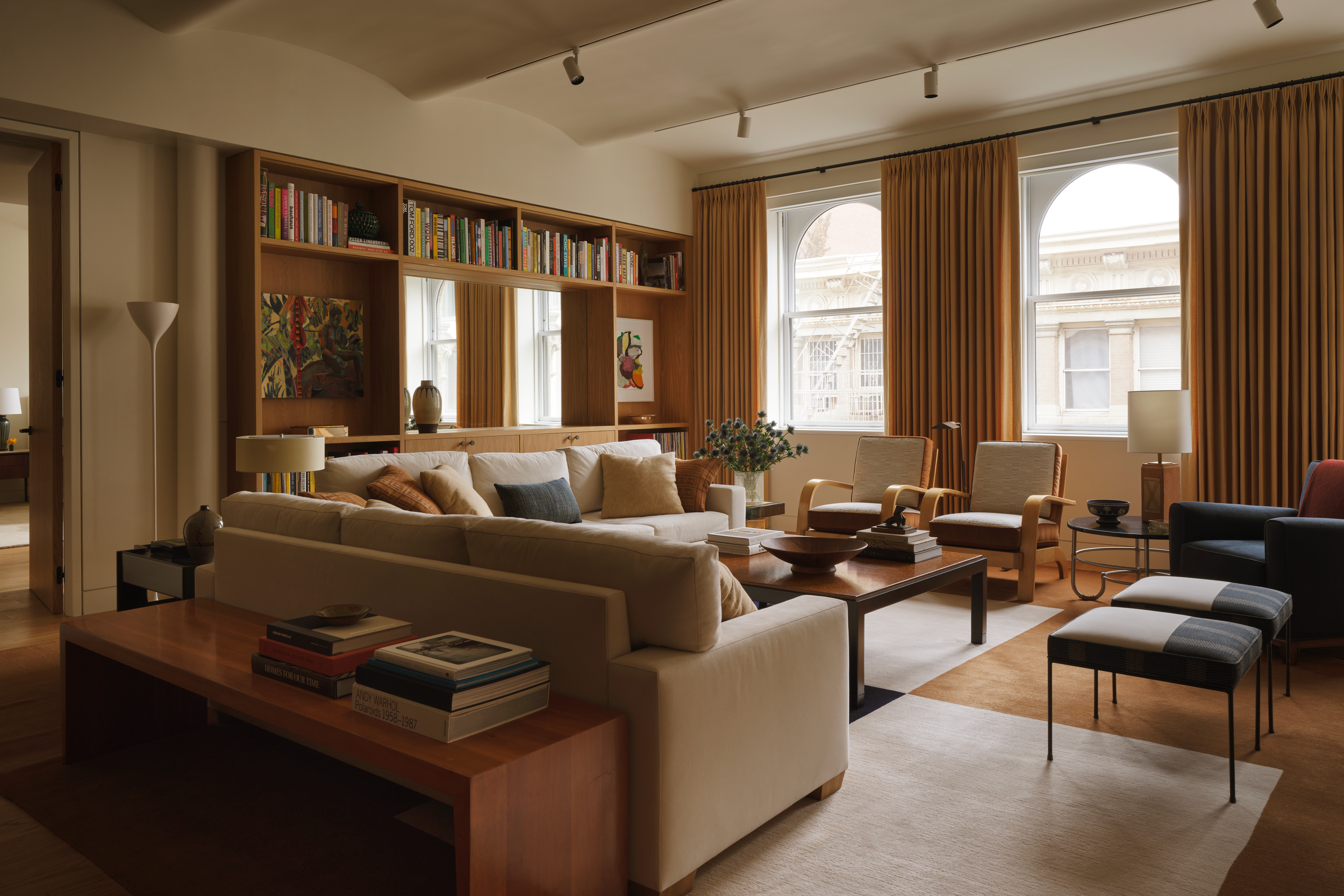 Living room with a beige sofa, armchairs, and a wooden coffee table, large windows with yellow curtains, bookshelves, and framed artwork.