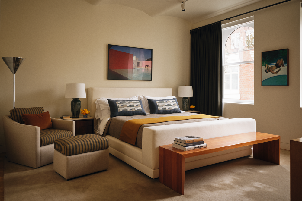 A modern bedroom with a large white bed, patterned pillows, yellow blanket, and wooden bench. There is a striped armchair with a pillow and ottoman on the left, side tables with lamps, colorful wall art, and windows with black curtains.