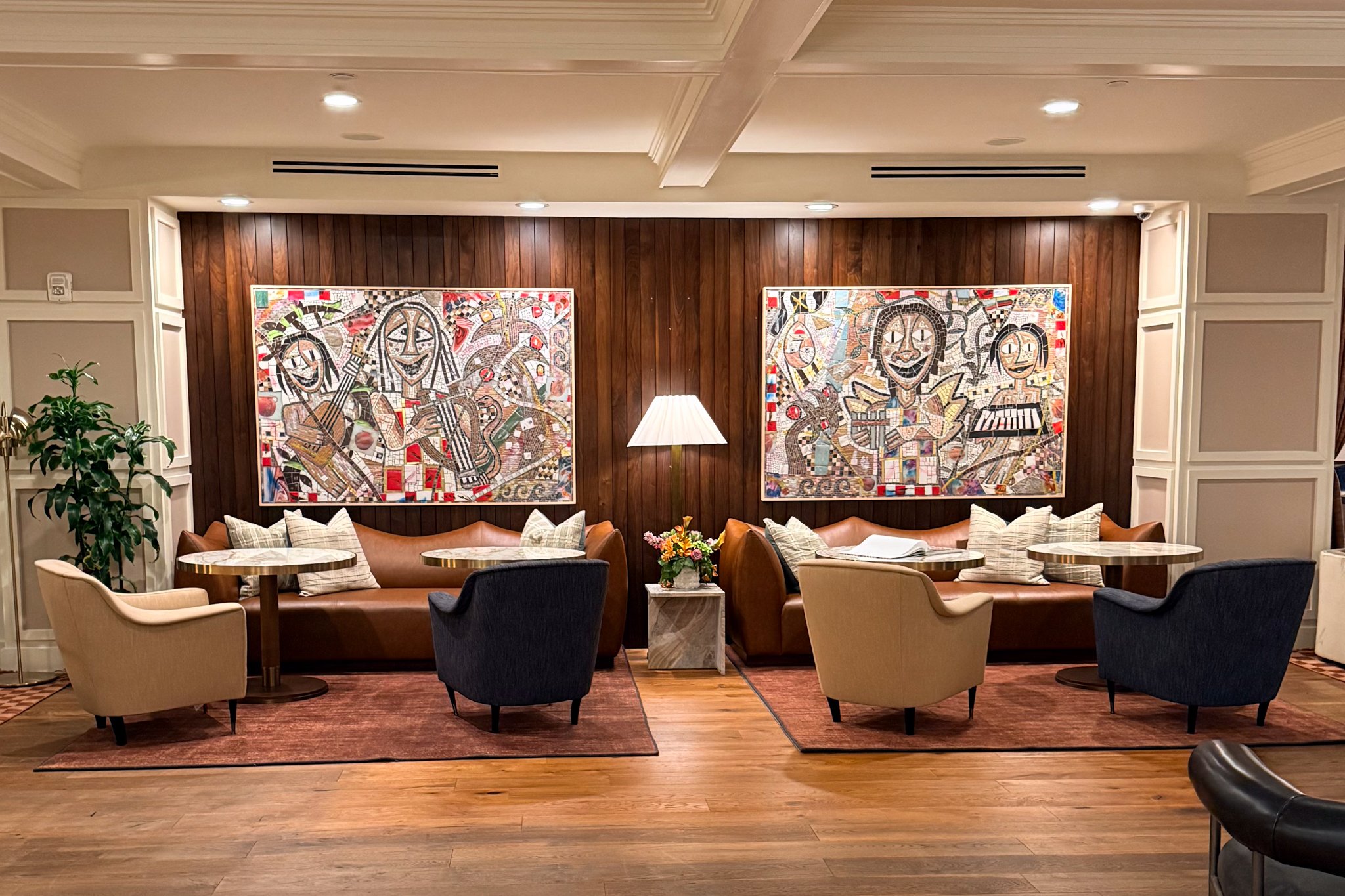 A cozy sitting area with two brown sofas, four beige armchairs, and two black armchairs, arranged around small round tables. A lamp and a flower arrangement are placed between the sofas, and abstract colorful artwork decorates the wooden-paneled wall