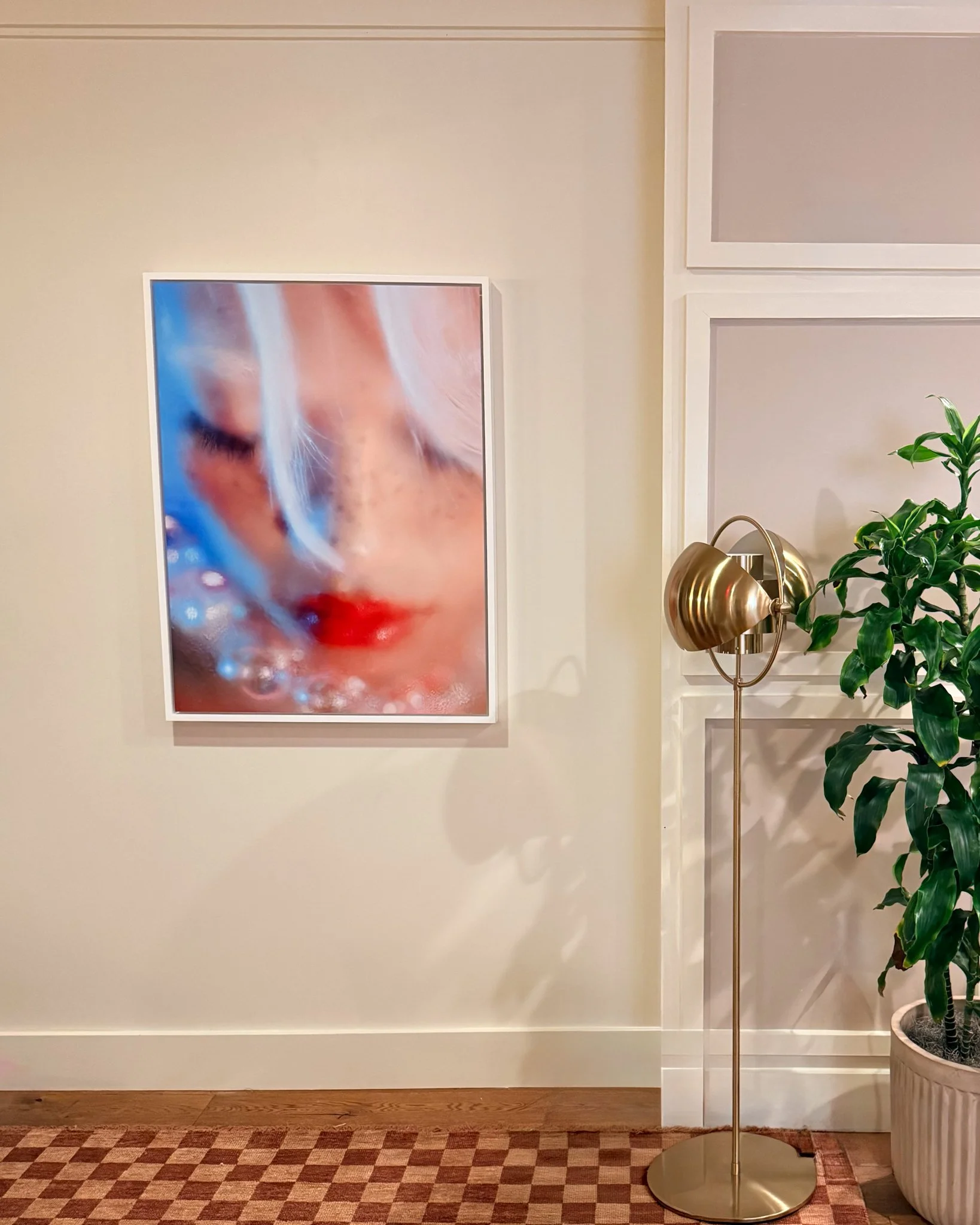 Photography of a woman's face with closed eyes, bright red lips, light hair, and a dreamy, blurred effect, hung on a beige wall beside a gold-colored floor lamp and a potted green plant.
