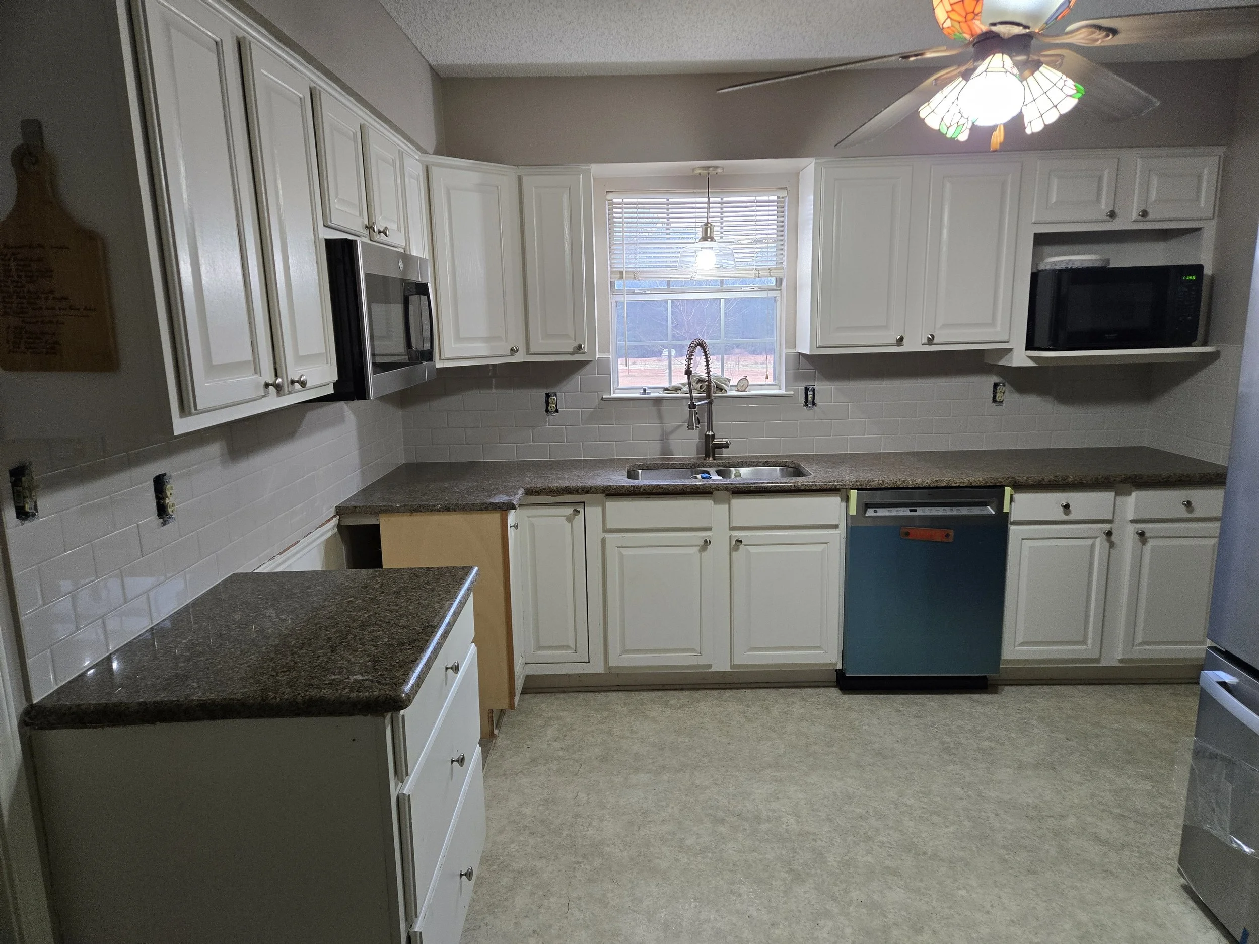 Empty kitchen with white cabinets, granite countertops, a window above the sink, a ceiling fan with colorful stained glass design, and appliances including a microwave, dishwasher, and refrigerator.