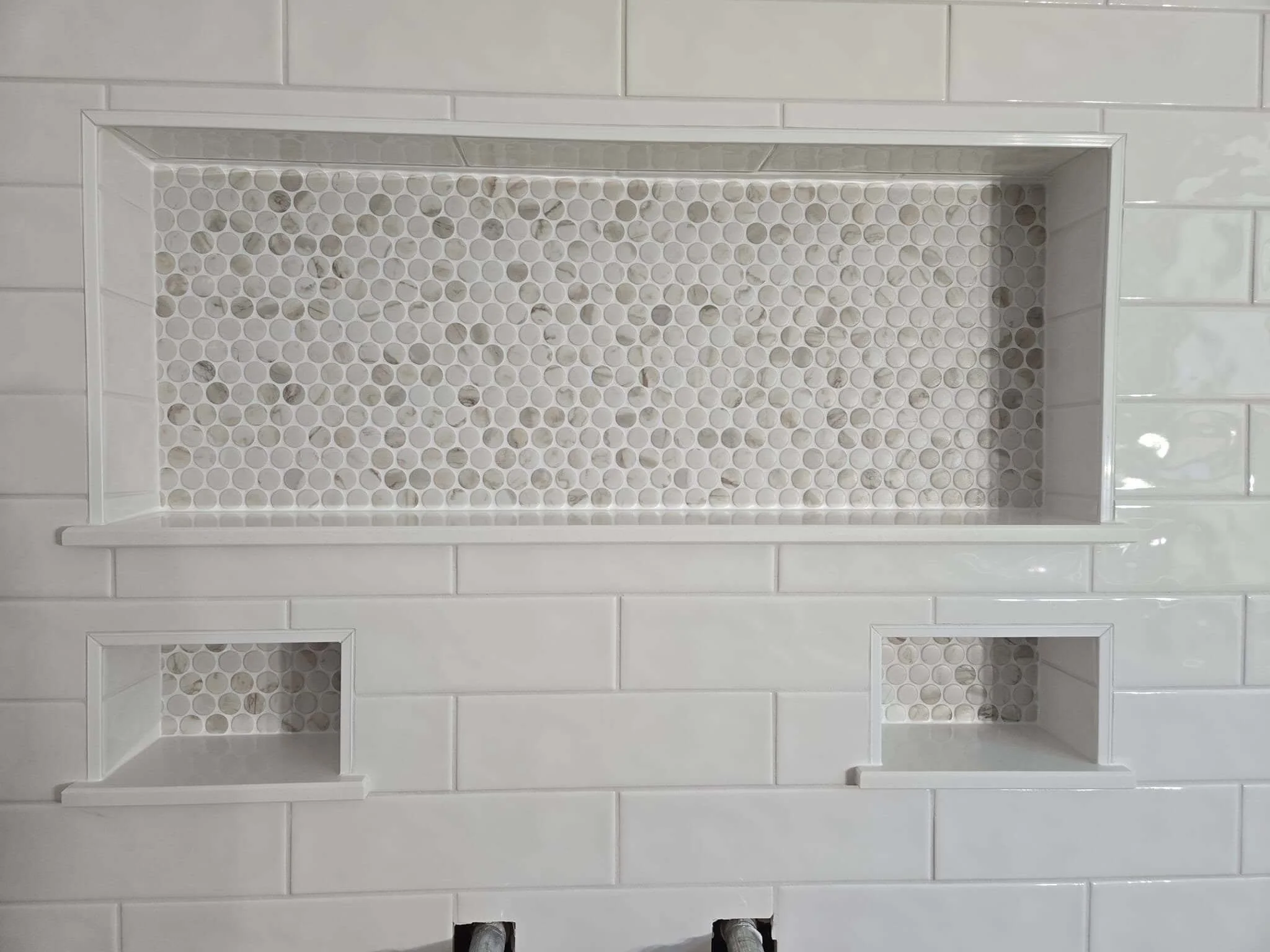 A wall with three recessed shelves: two small square ones with a mosaic tile pattern inside, and one larger rectangular one with a similar mosaic pattern. The wall is tiled with white, rectangular subway tiles.