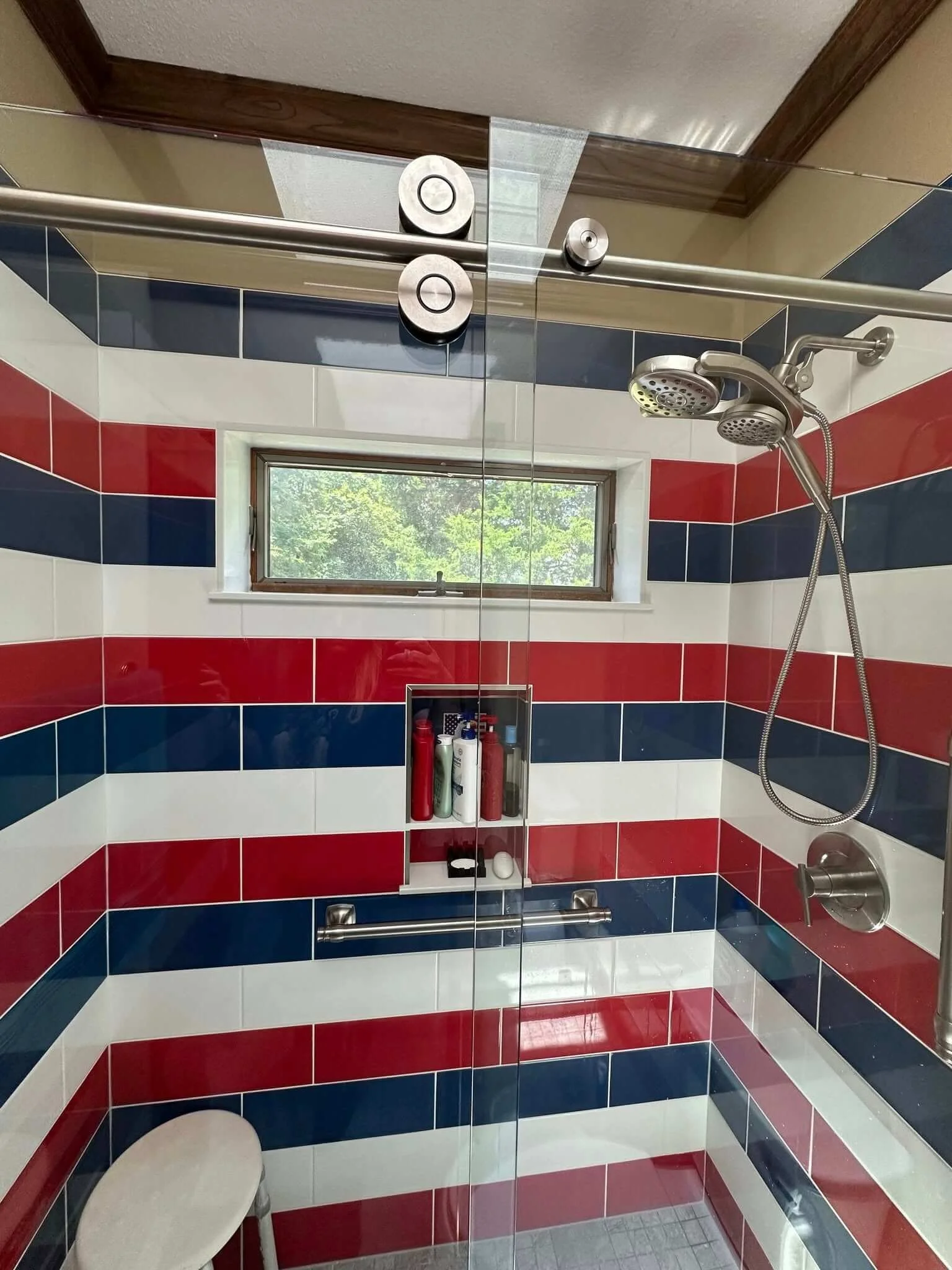 Shower with a red, white, and blue stripe tiled wall, small window, glass door, and showerhead, with a built-in niche holding shampoo and soap.
