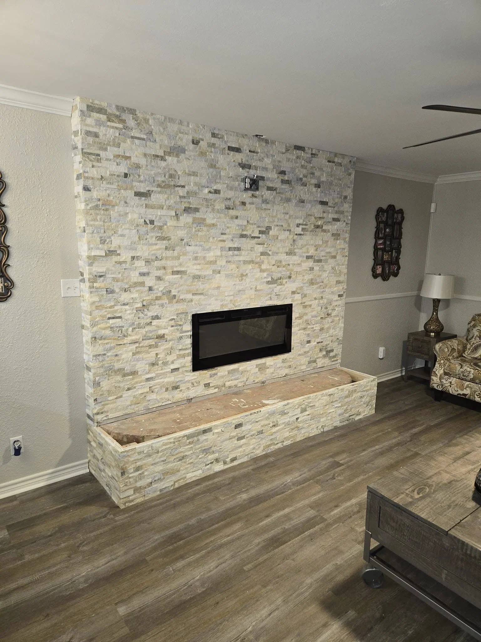 Living room with a stone fireplace and a wooden mantel, beige sofa with floral pattern, side table with lamp, and wood flooring.