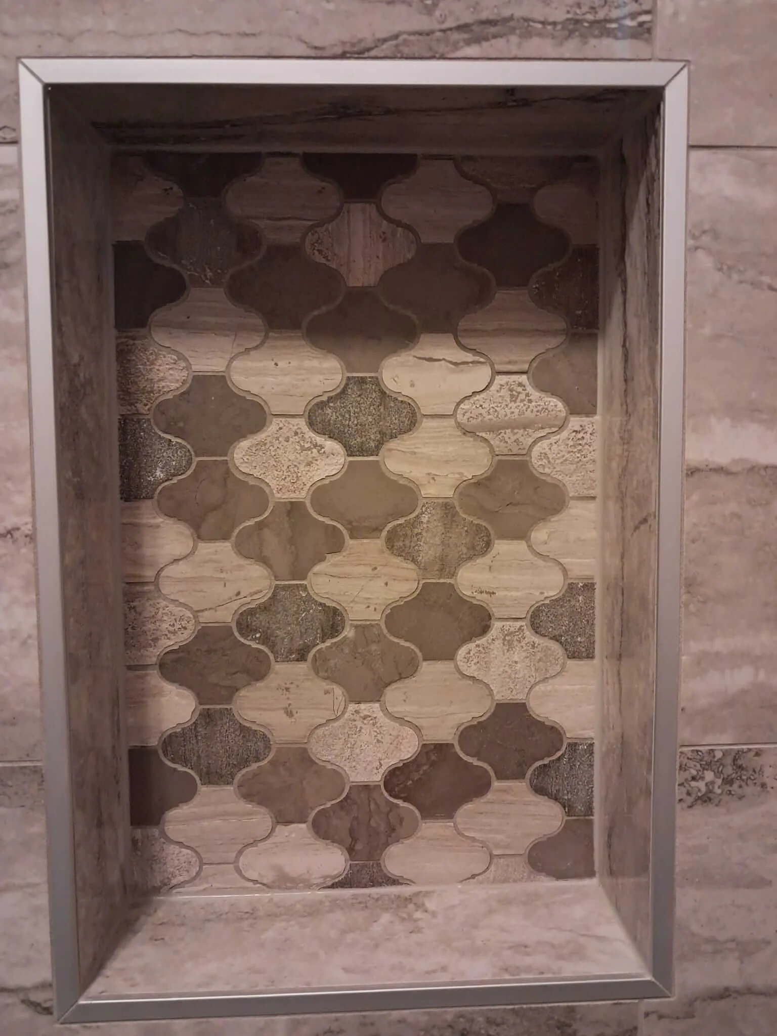 Recessed wall niche with a decorative inlay of patterned tiles in shades of brown and beige, framed by metal trim, on a stone wall.