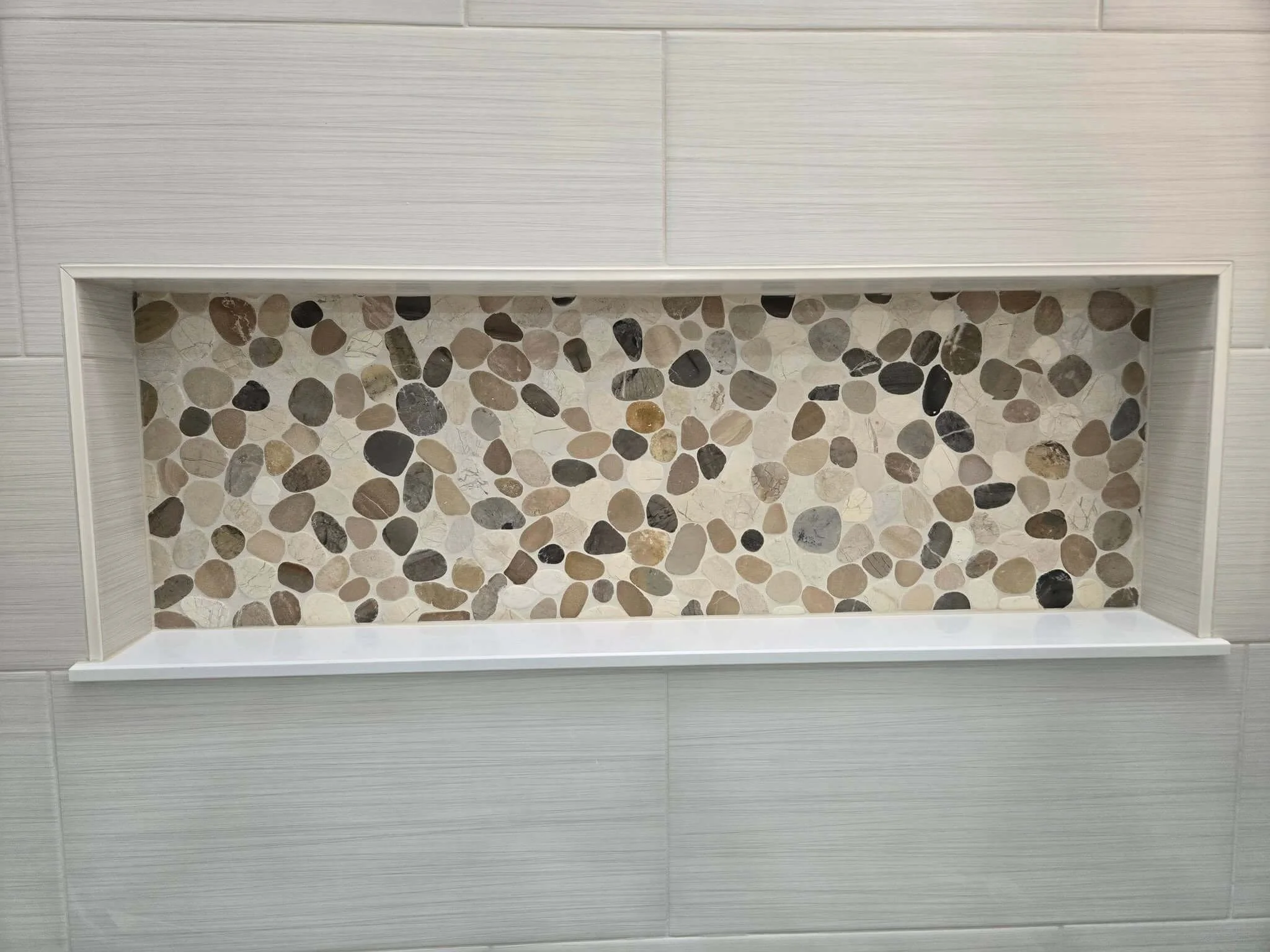 A rectangular wall niche with a mosaic pebble interior and a white border, embedded in a light-colored tiled wall.