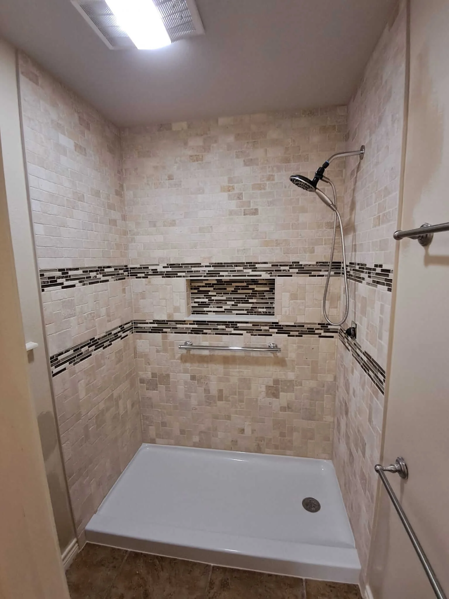 Shower with beige and brown tiled walls, black and brown decorative tile accents, a showerhead, a grab bar, and a white shower base.