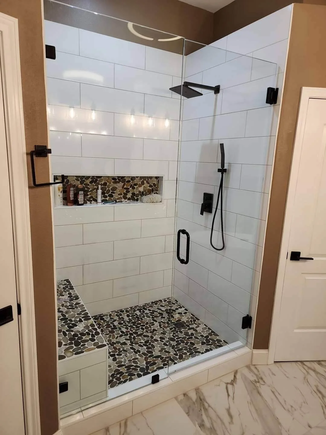 Modern walk-in shower with white subway tiles, pebble mosaic floor, built-in niche with toiletries, black fixtures, and a glass enclosure.