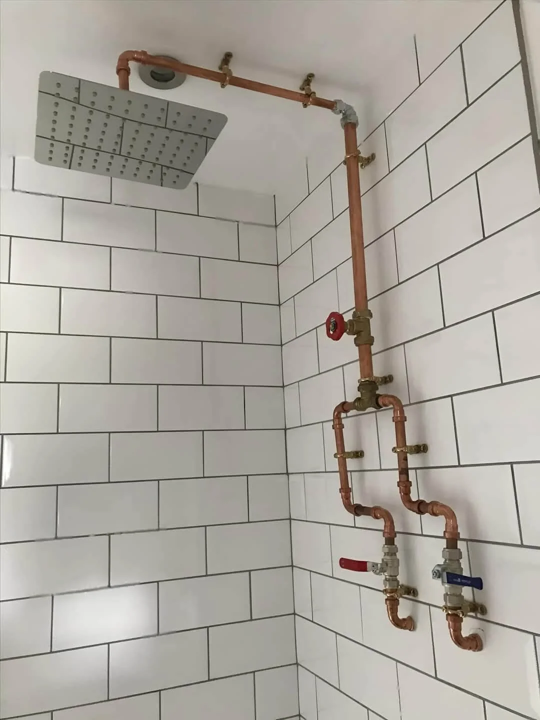 Image of a showerhead and plumbing pipes installed on white tiled wall in a bathroom.