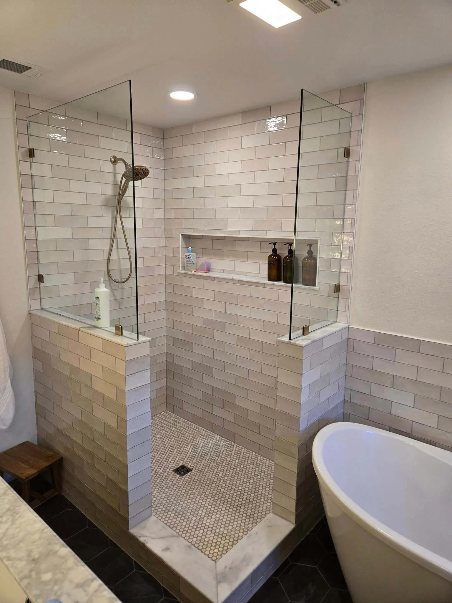 Modern bathroom with a walk-in shower enclosed by glass panels, beige subway tile walls, a niche with toiletries, and a freestanding bathtub nearby.
