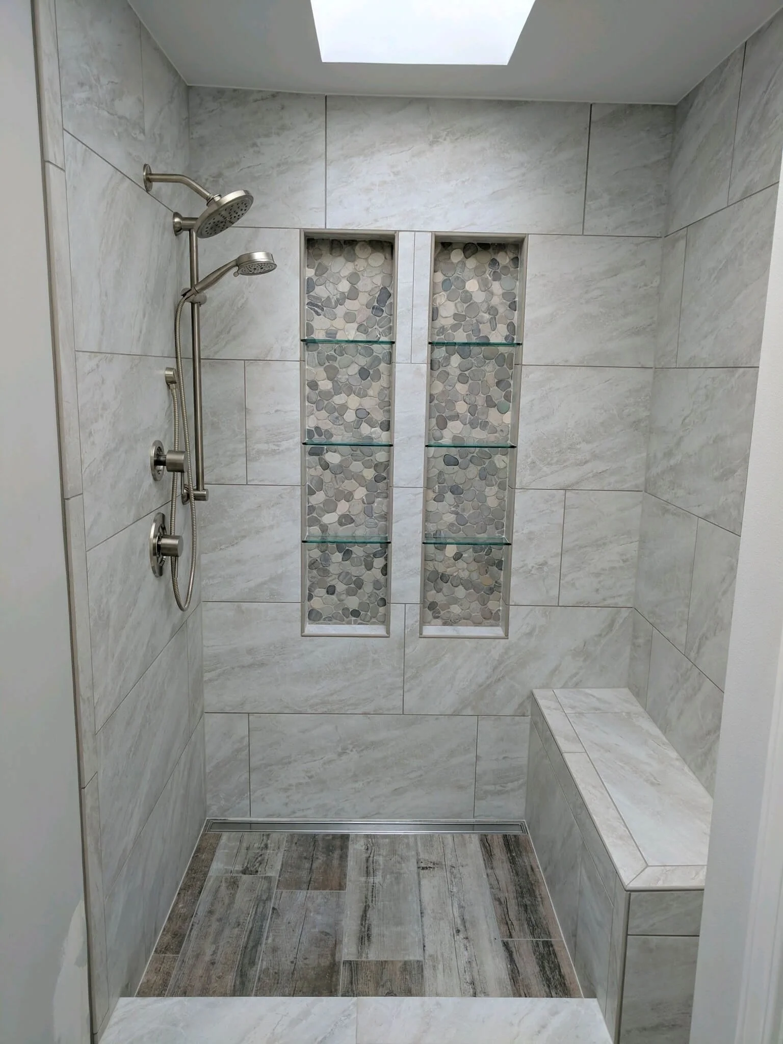 Walk-in shower with light gray marble tiles, a built-in tiled bench, and two vertical recess shelves with pebble stone backgrounds and glass shelves.