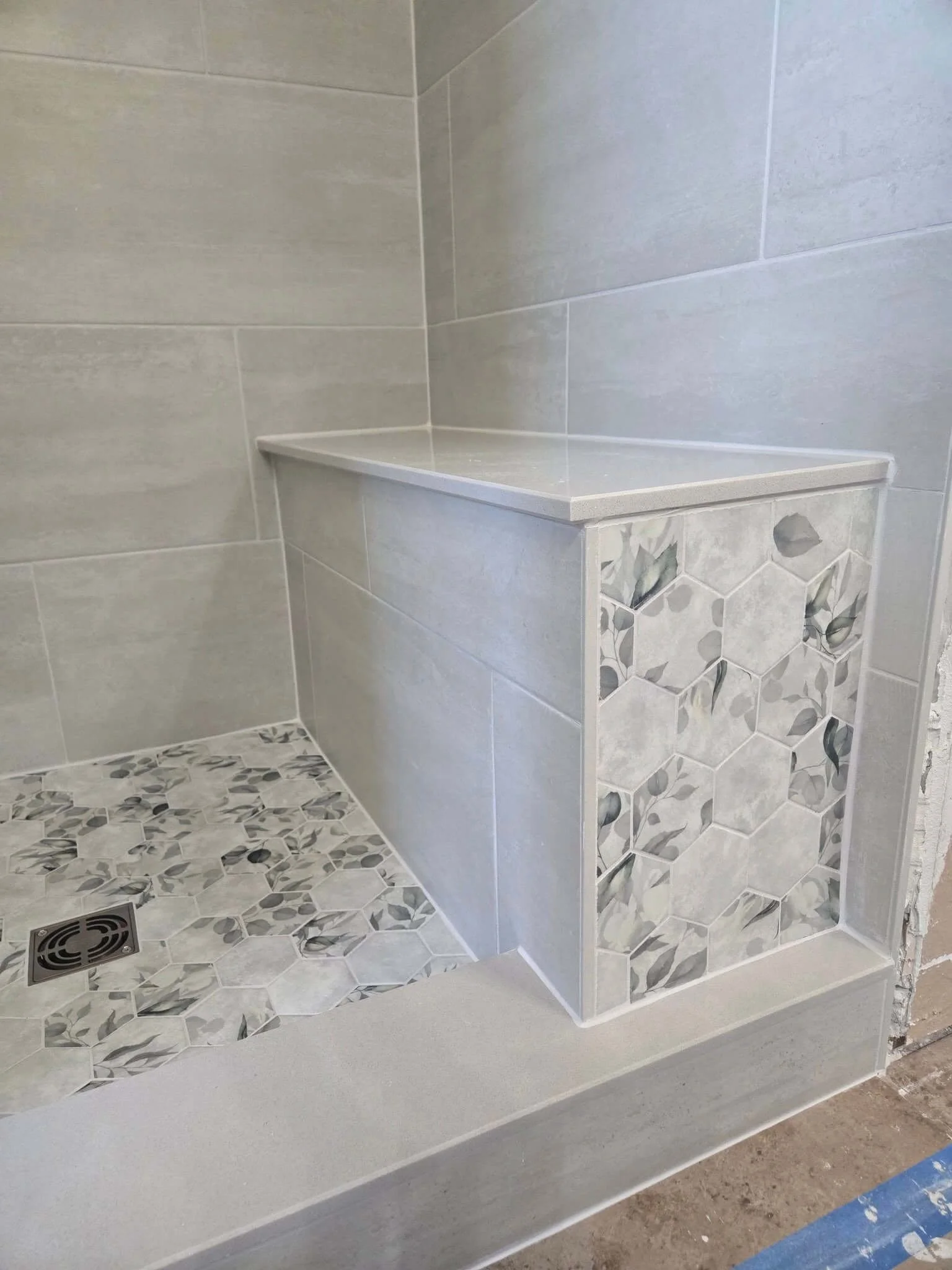 A corner of a tiled shower with a built-in shelf, gray wall tiles, and a patterned accent tile on the side of the shelf, featuring hexagonal and floral motifs.
