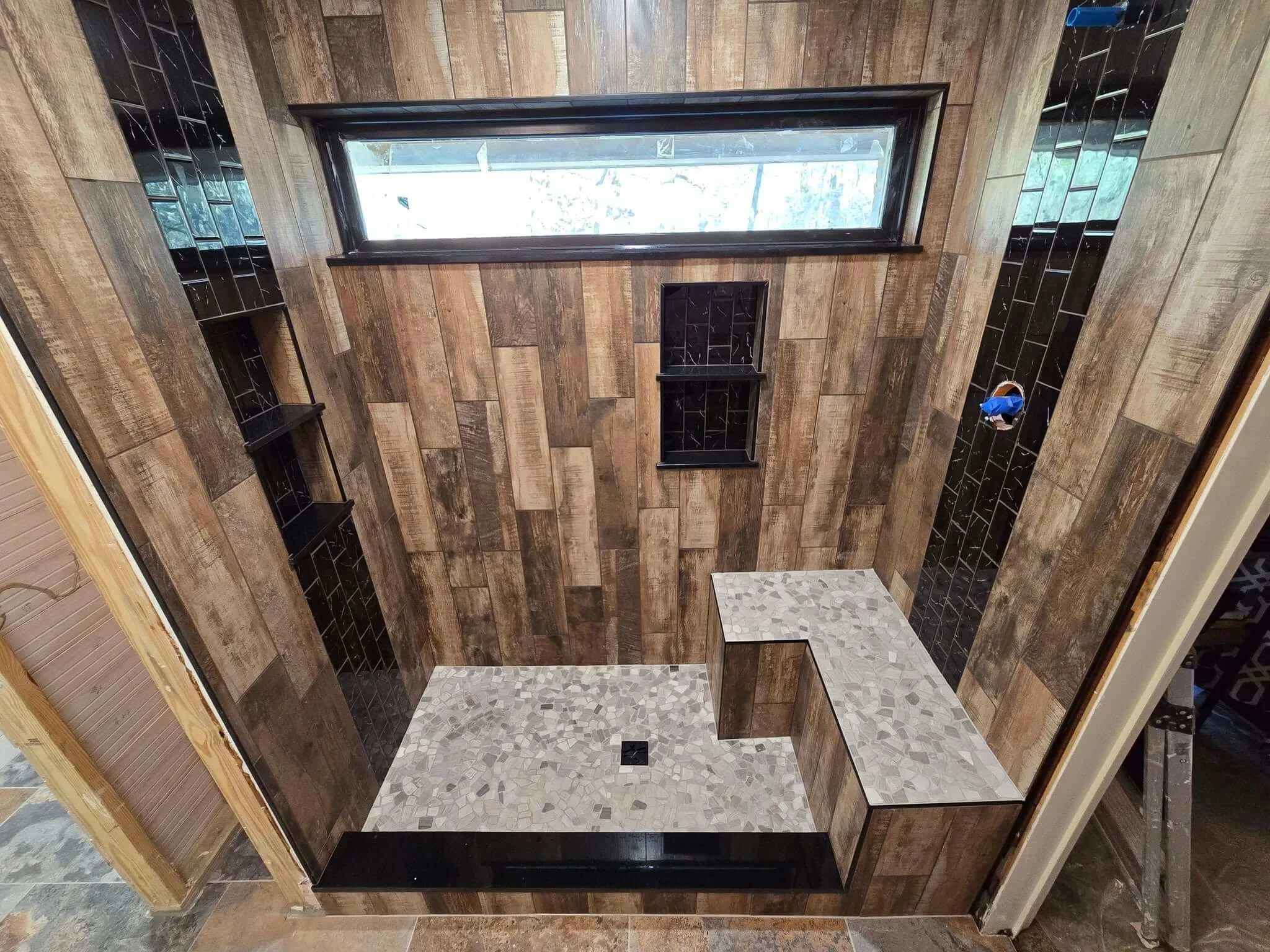 A modern shower with wooden wall panels, black tile accents, a window, built-in shelves, and a pebble-textured bench and floor.