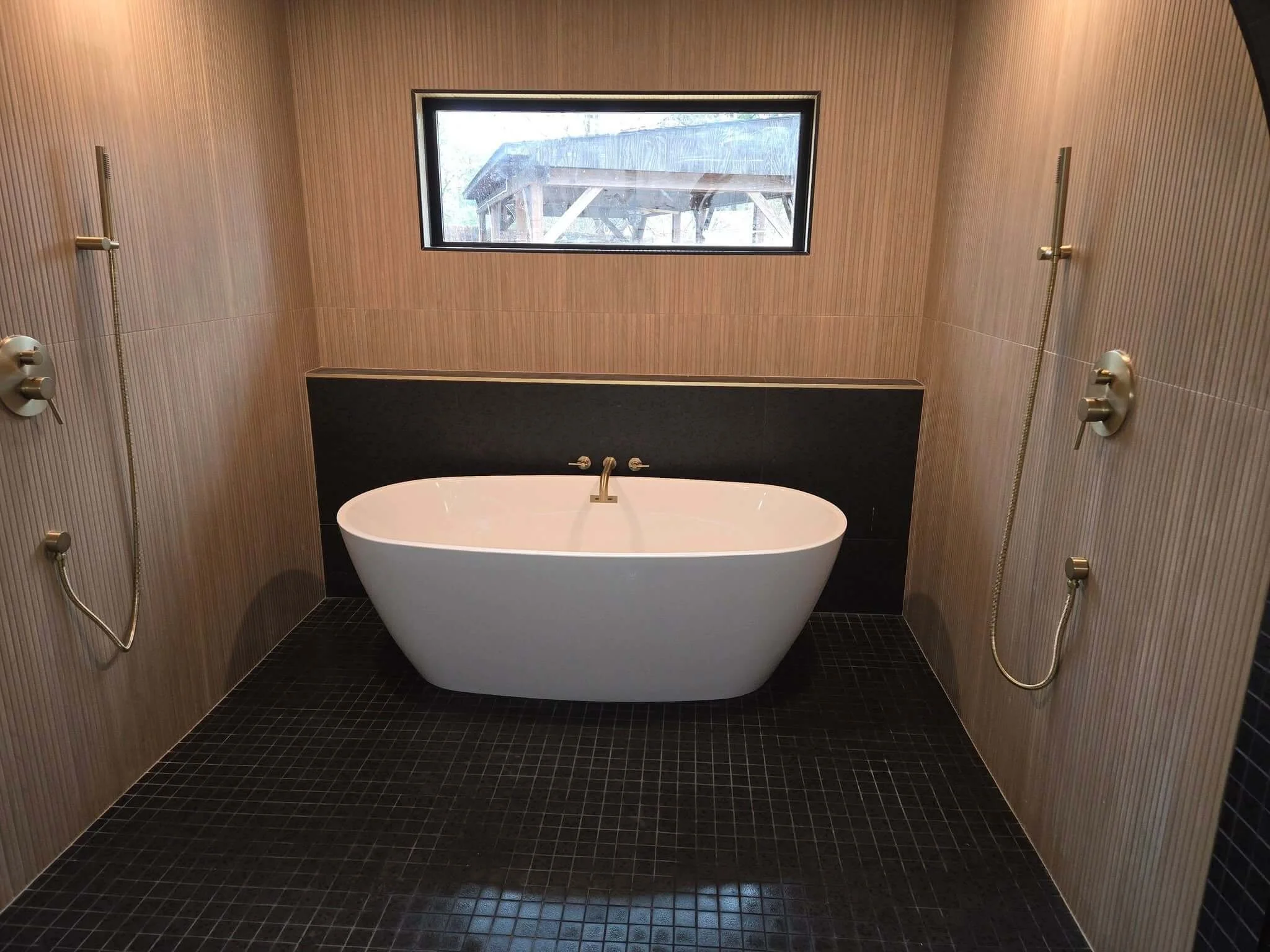 Modern bathroom with a free-standing white bathtub, black tiled floor, wood-paneled walls, and a rectangular window showing a wooden structure outside.
