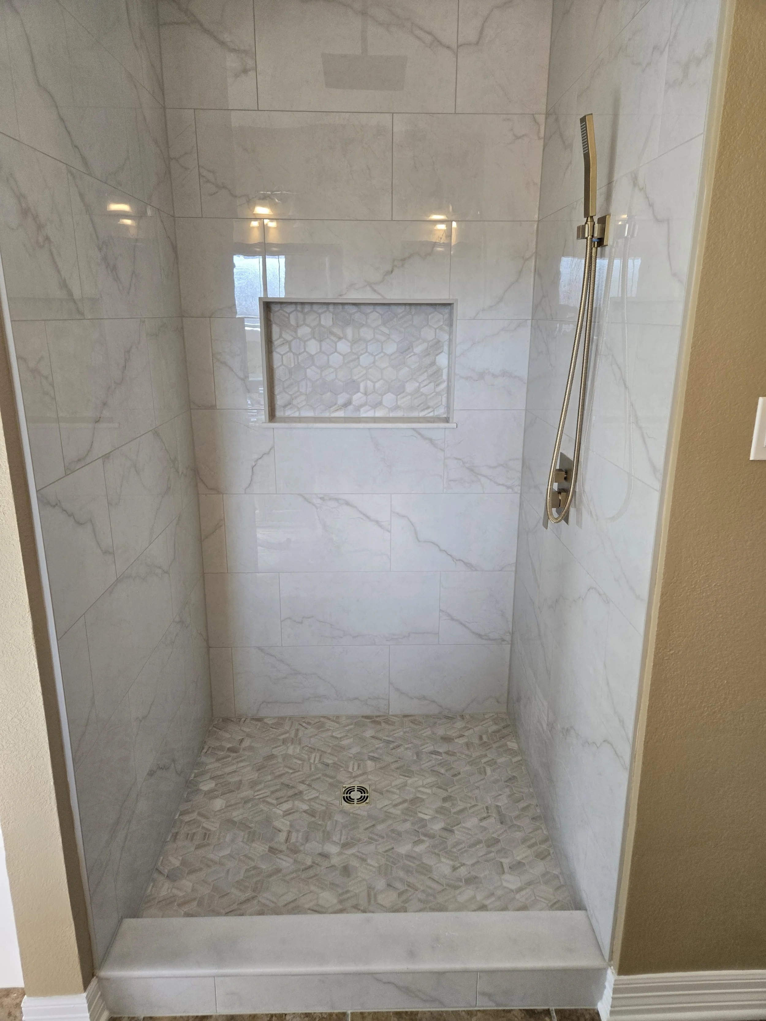 A modern walk-in shower with white marble tiles on the walls, a recessed niche with hexagonal tiles, a handheld showerhead with a flexible hose, and a drain on the floor.