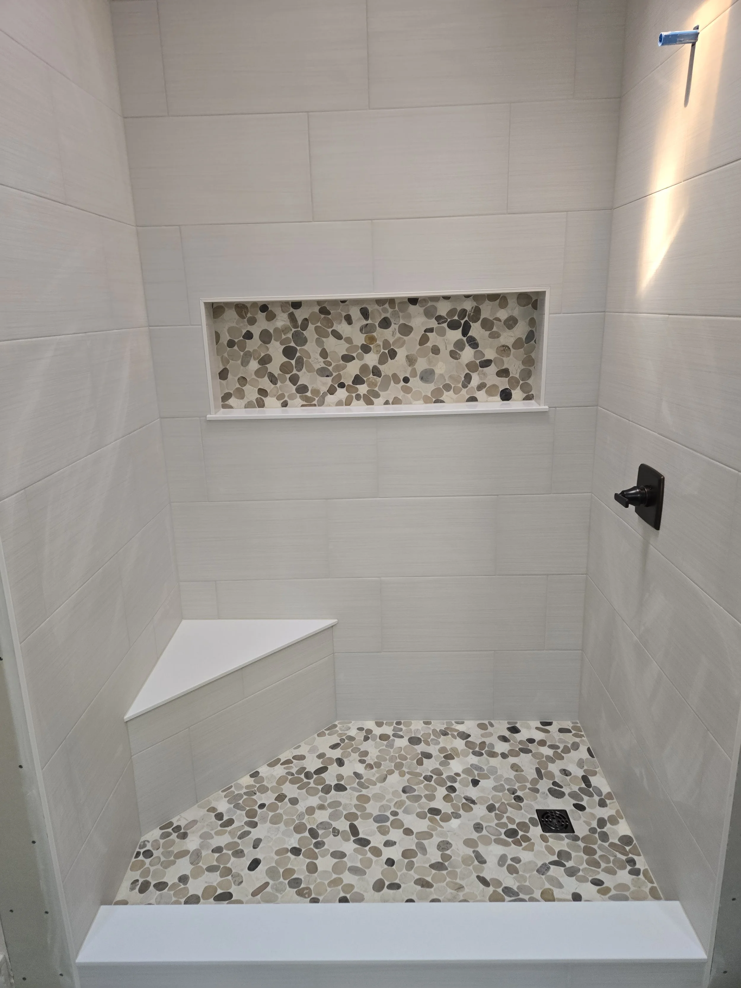 Empty shower with beige tiled walls, pebble mosaic floor and a built-in corner seat. There is a horizontal pebble mosaic niche on the wall and a showerhead on the upper right.