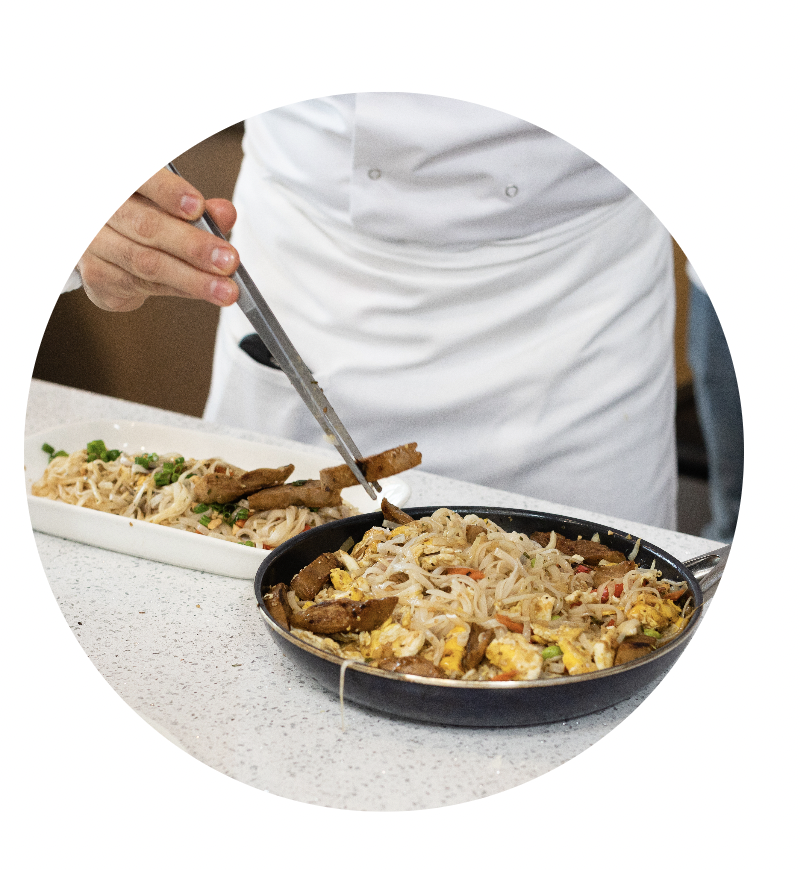 A chef serving stir-fried noodles with vegetables and meat onto a white rectangular plate from a black frying pan.