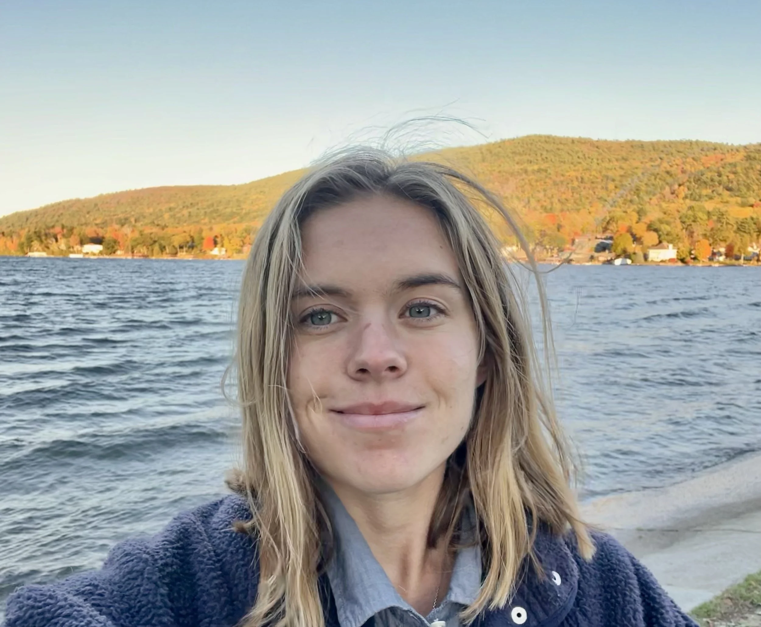 A woman with shoulder-length blonde hair taking a selfie at a lake during autumn, with rolling hills covered in colorful fall foliage in the background.
