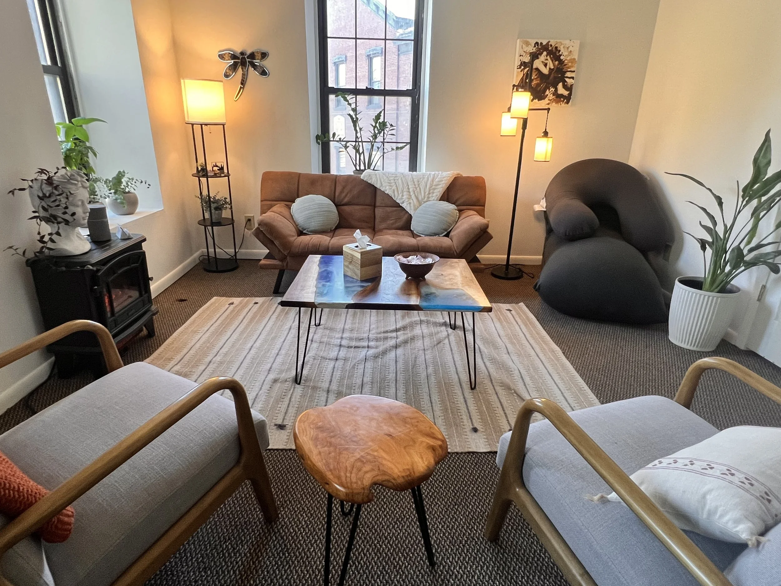 A cozy room with a brown sofa, a black lounge chair, two armchairs, a wooden coffee table, a small wooden side table, a floor lamp, a statue, potted plants, and wall art, with natural light coming through large windows.